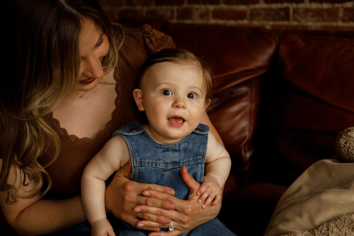 A smiling baby in denim overalls sits on a mother's lap near a brown leather couch in warm indoor lighting.