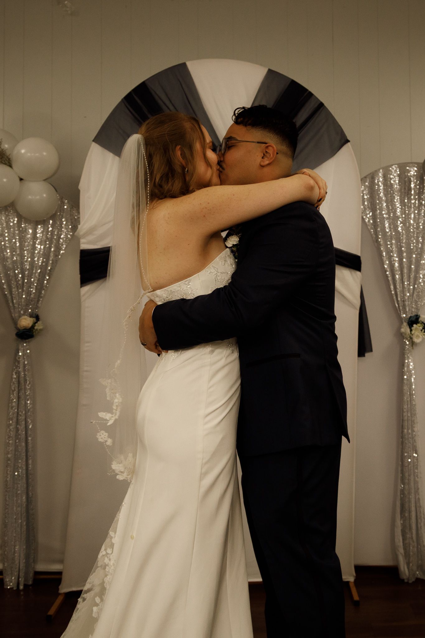 A romantic wedding kiss under an archway decorated with silver sparkle curtains and white balloons.