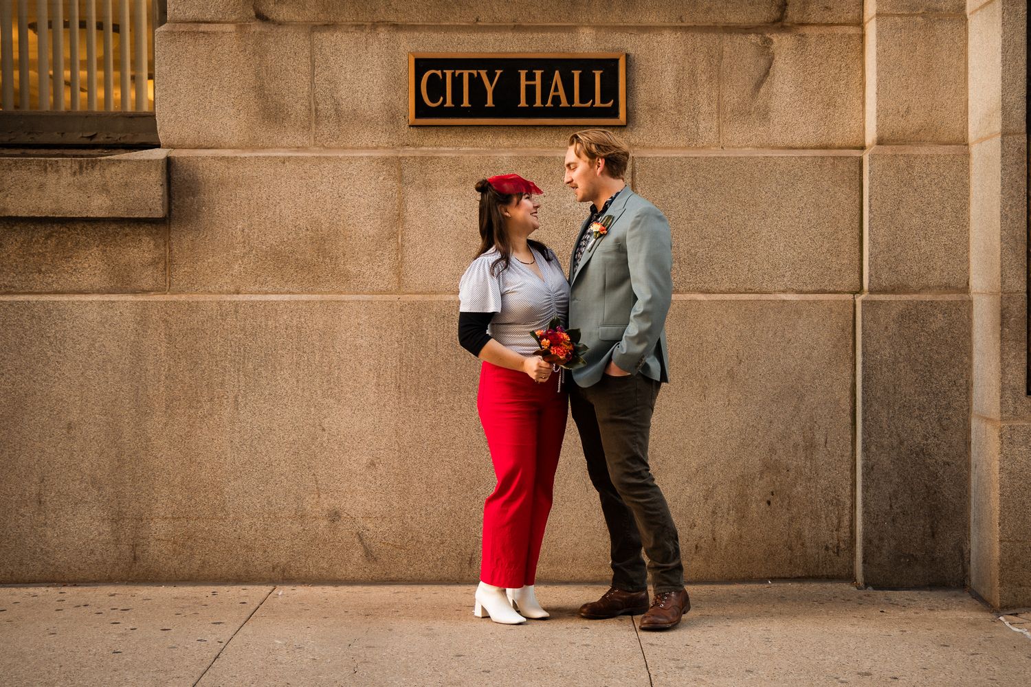 A couple in red and grey attire poses for photos outside a city hall building.