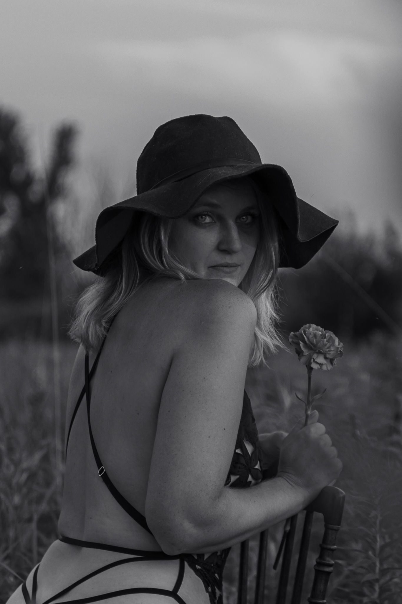 Black and white portrait of person in wide brim hat holding flower in field.