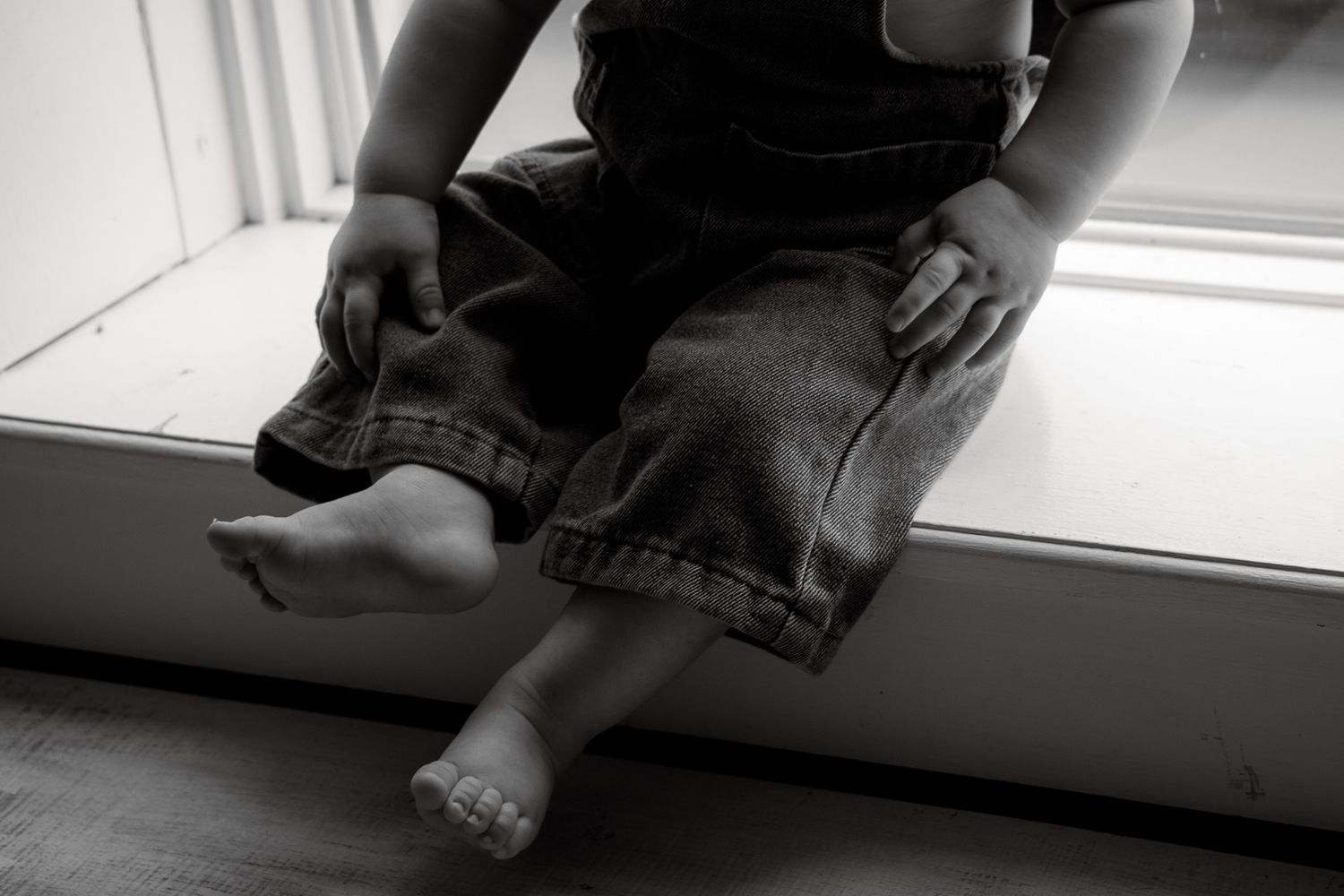 Black and white photo of a young child sitting on a windowsill, wearing denim overalls with bare feet dangling.