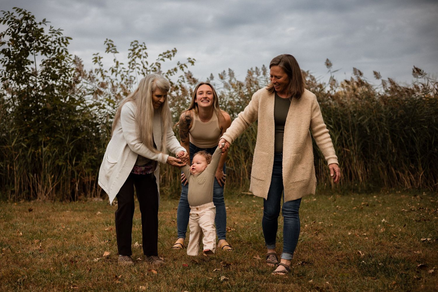 Three people in cozy sweaters share a joyful moment together in a field with tall grass on an overcast autumn day.