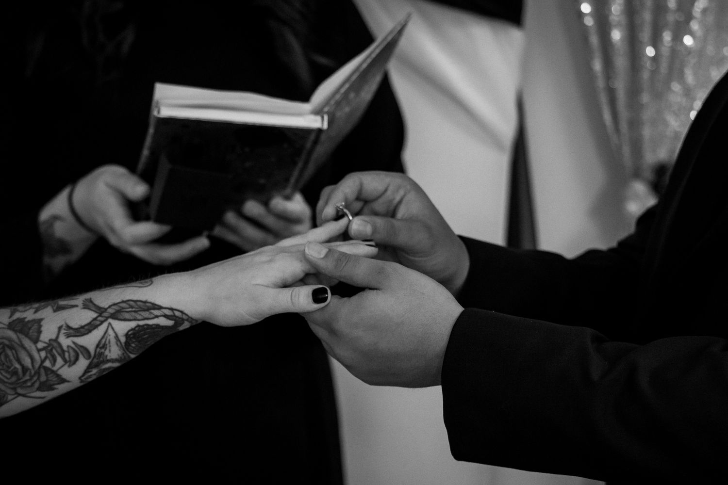 Black and white photo of hands exchanging wedding rings during a ceremony with someone holding a book in background.