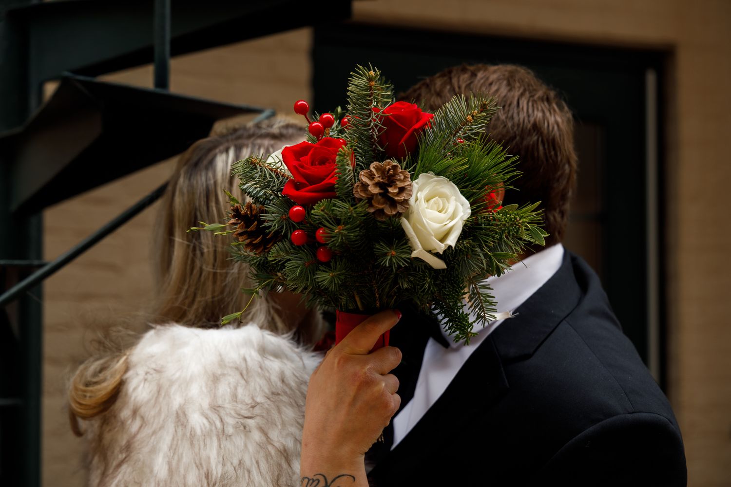A couple shares a romantic kiss during their winter wedding ceremony with red roses and evergreen decor.