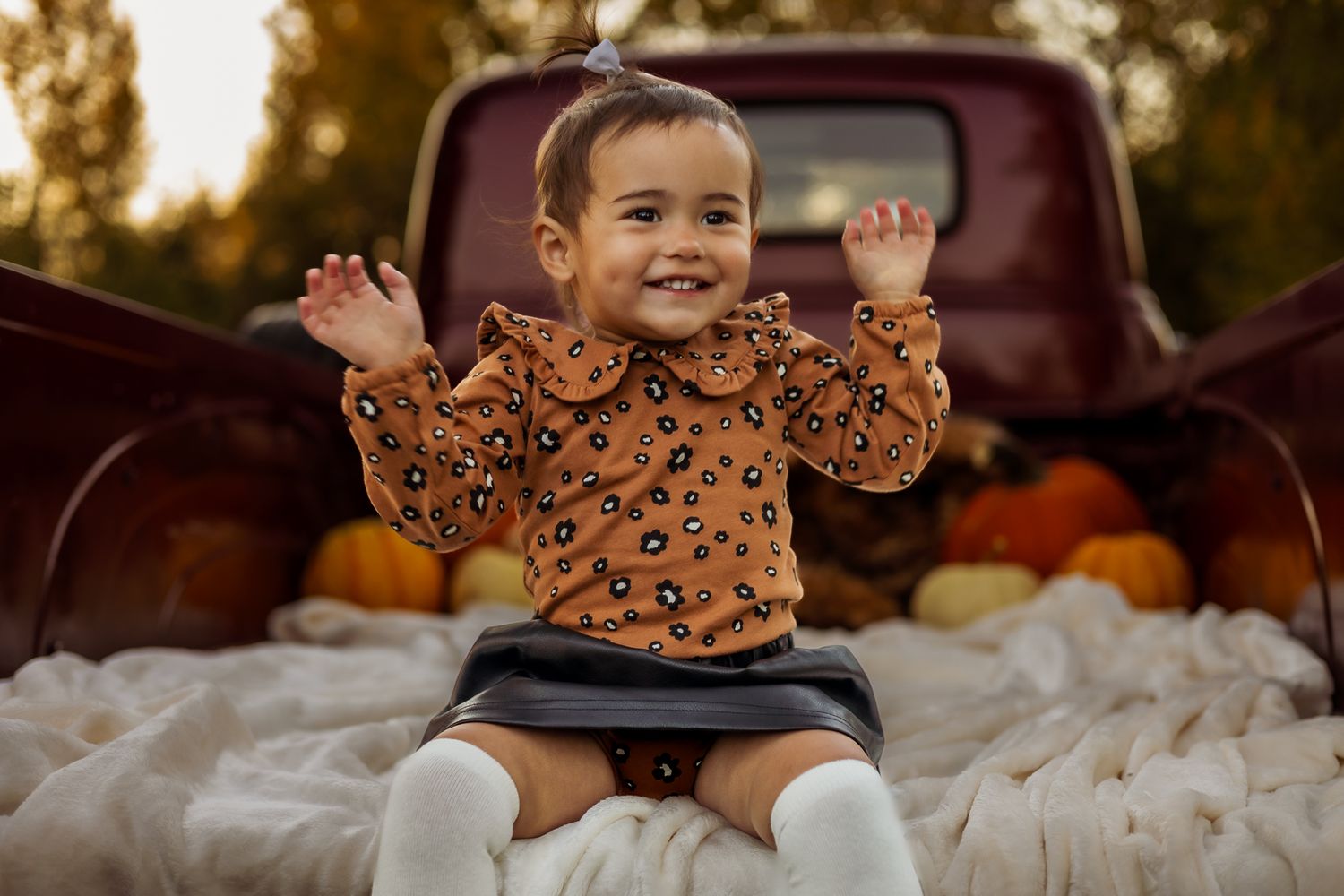 Small child wearing orange Halloween sweater sits in vintage truck bed filled with pumpkins and blankets at sunset.