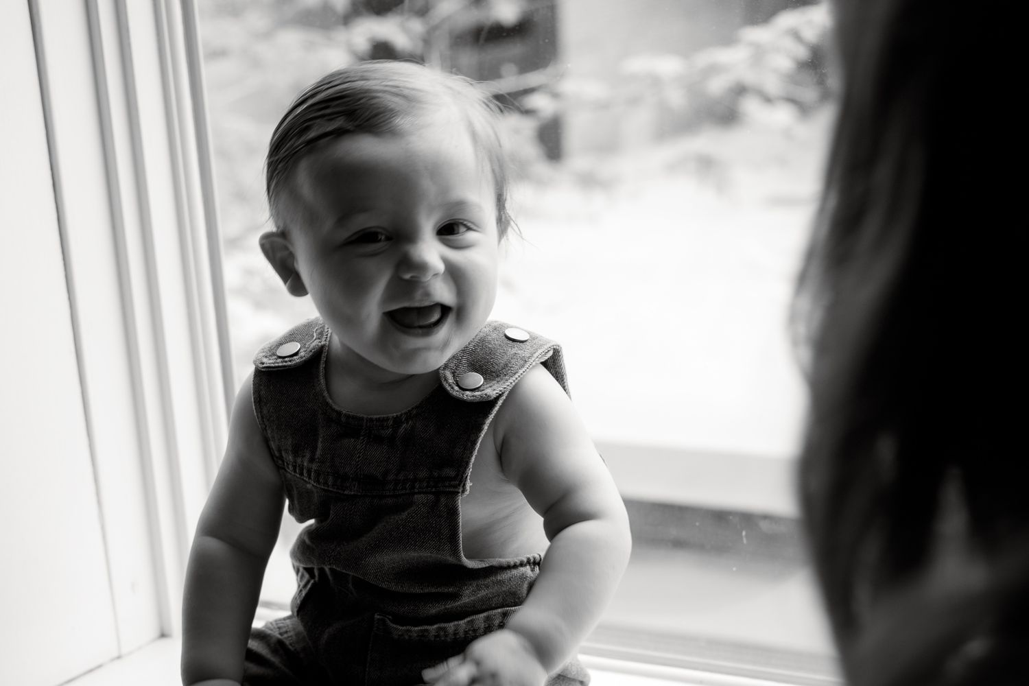 Black and white photo of a smiling baby in overalls sitting by a window with soft natural light.