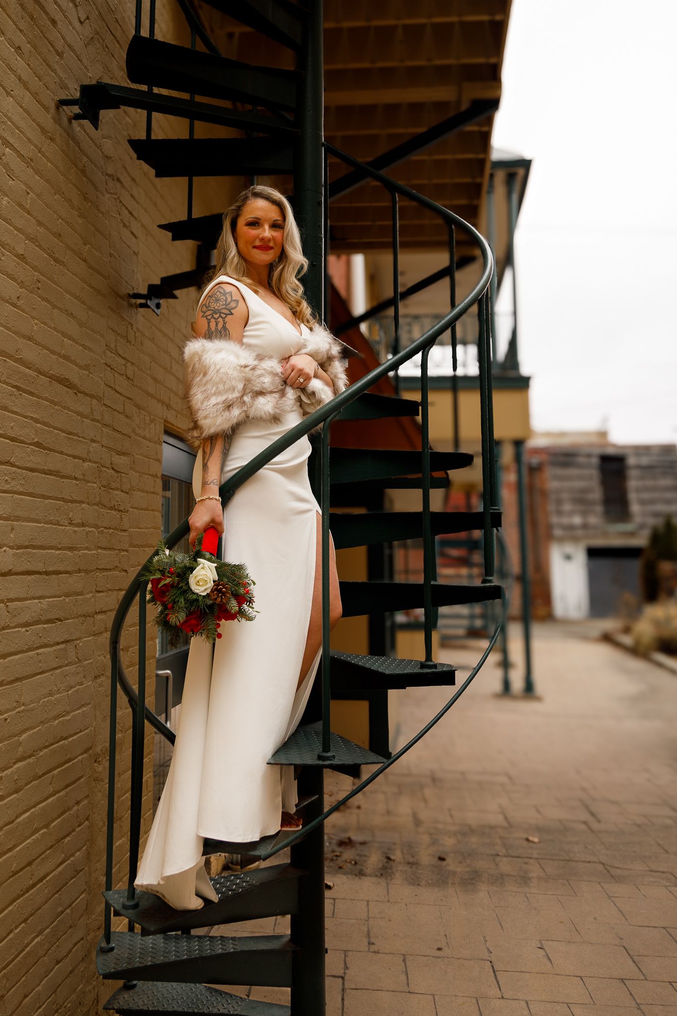 A bride in a white winter coat ascends a black spiral staircase while holding a bouquet of flowers.
