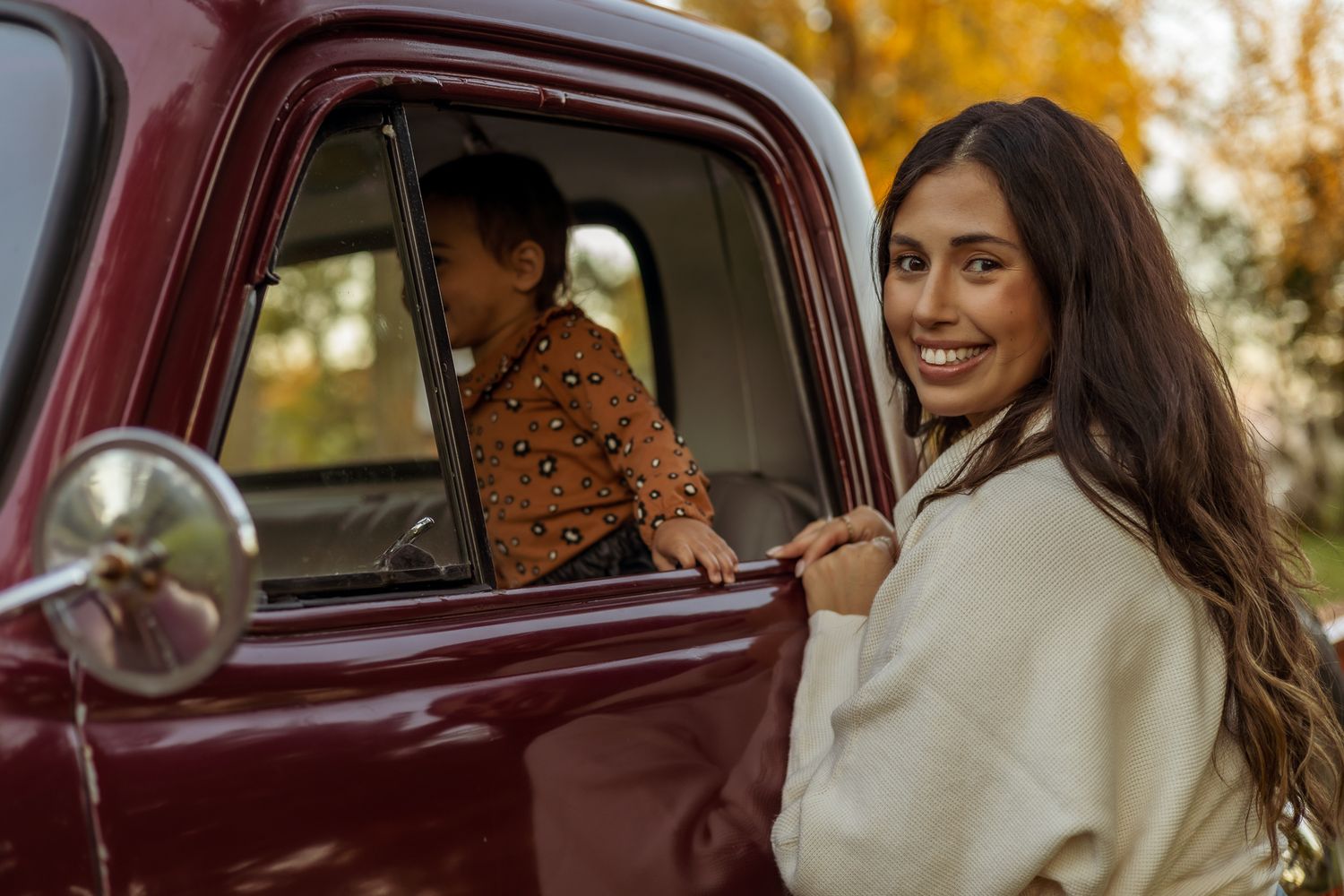A person sits in a burgundy vintage car during golden hour with autumn trees in the background.