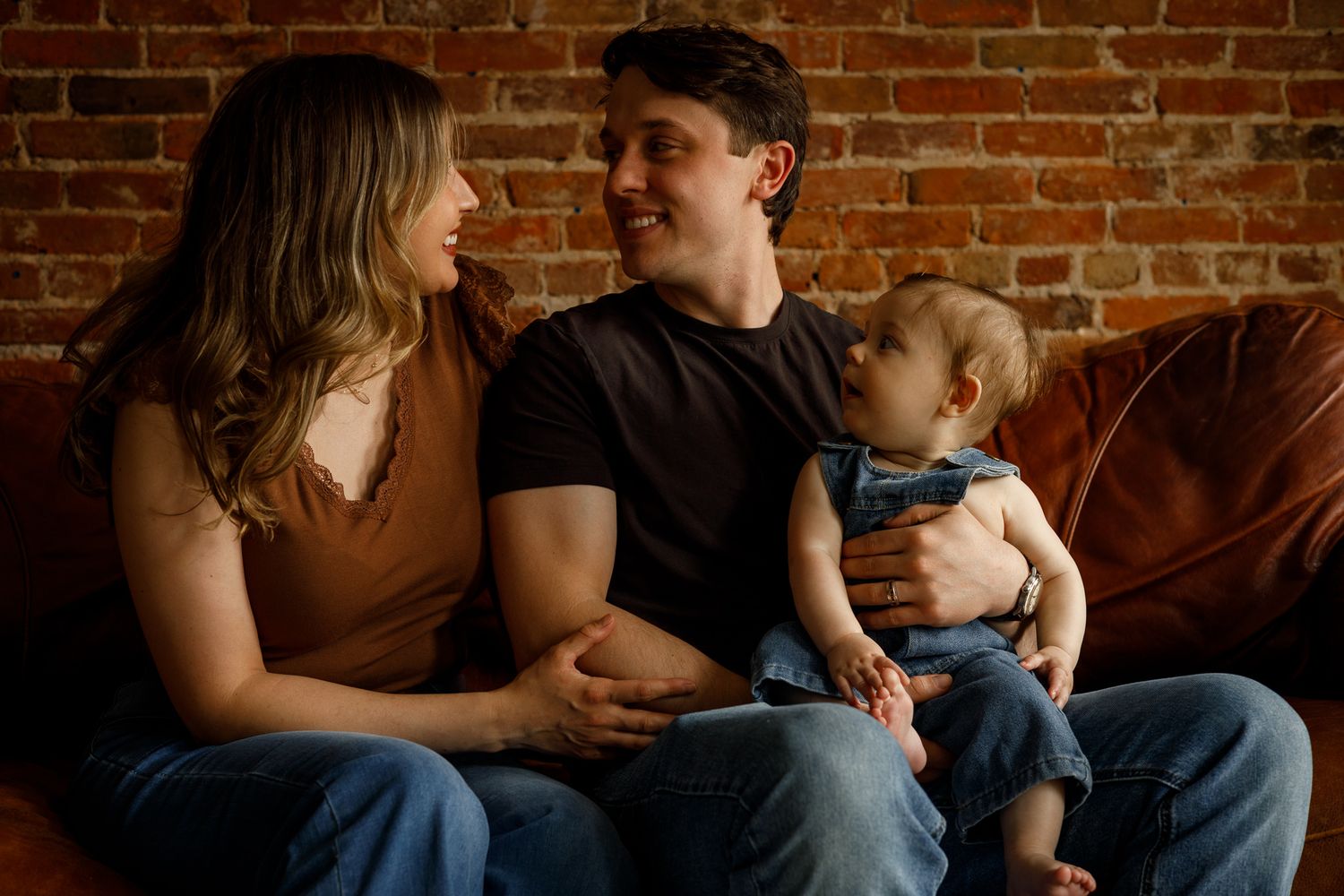 Young couple sitting on leather sofa smiling at each other while holding their baby against brick wall background.
