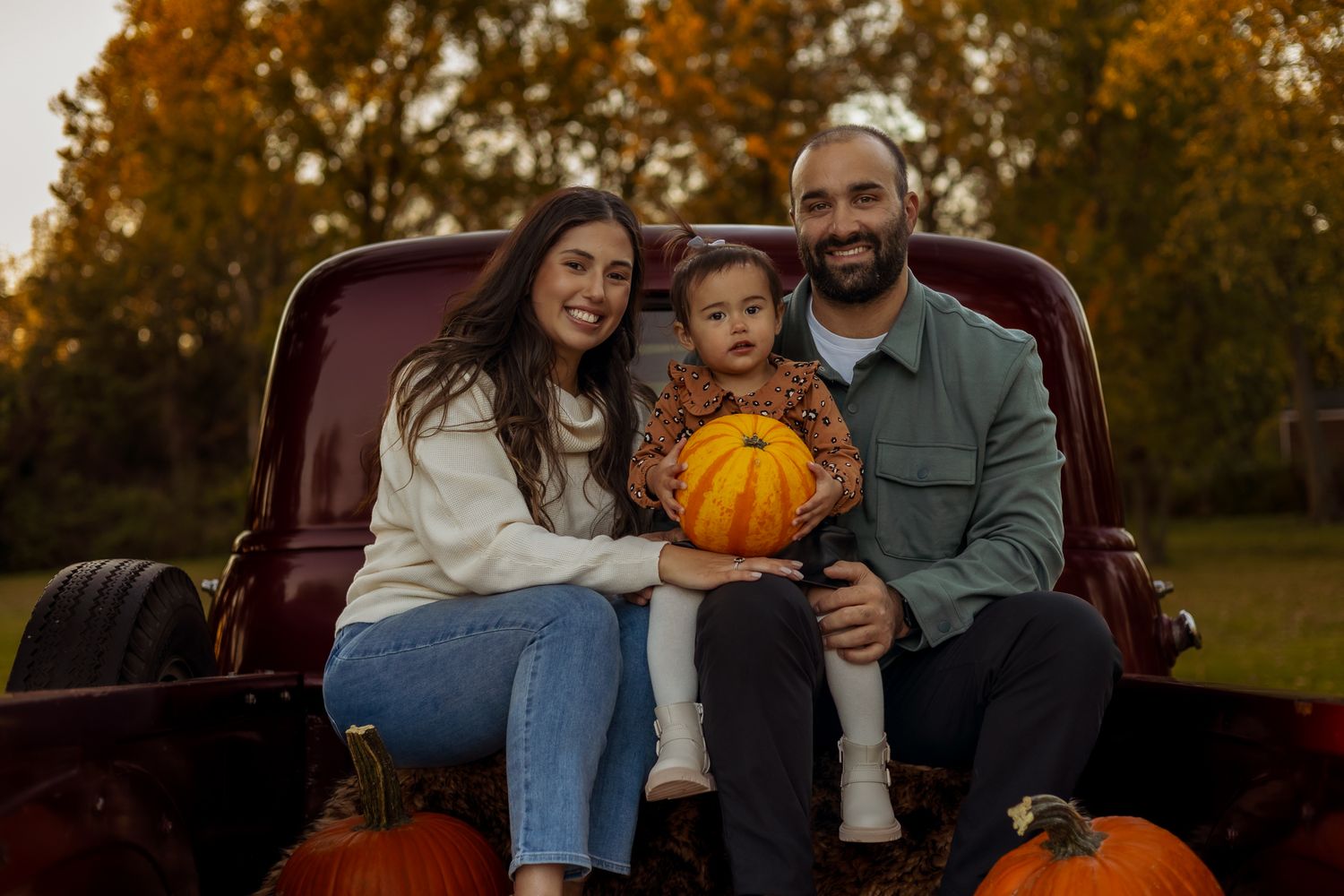 A family sits together on a vintage red truck during an autumn photoshoot with pumpkins at sunset.