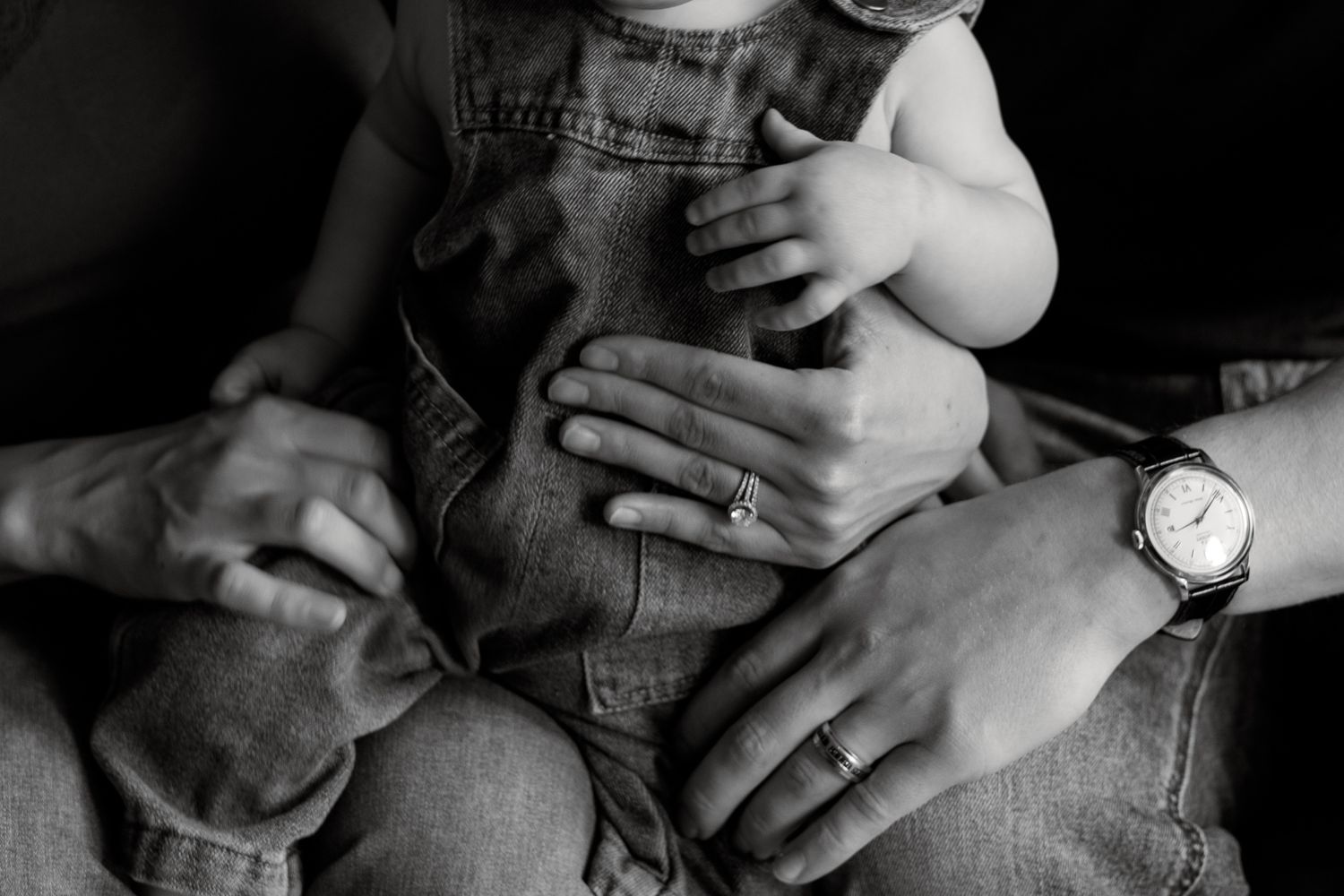 Black and white photo of parents holding a baby in denim overalls, showing a wedding ring and watch.