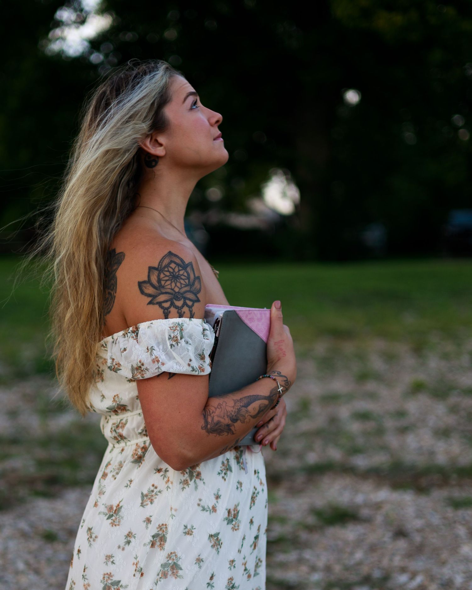 Woman in floral dress with lotus tattoo holding book outdoors at sunset.