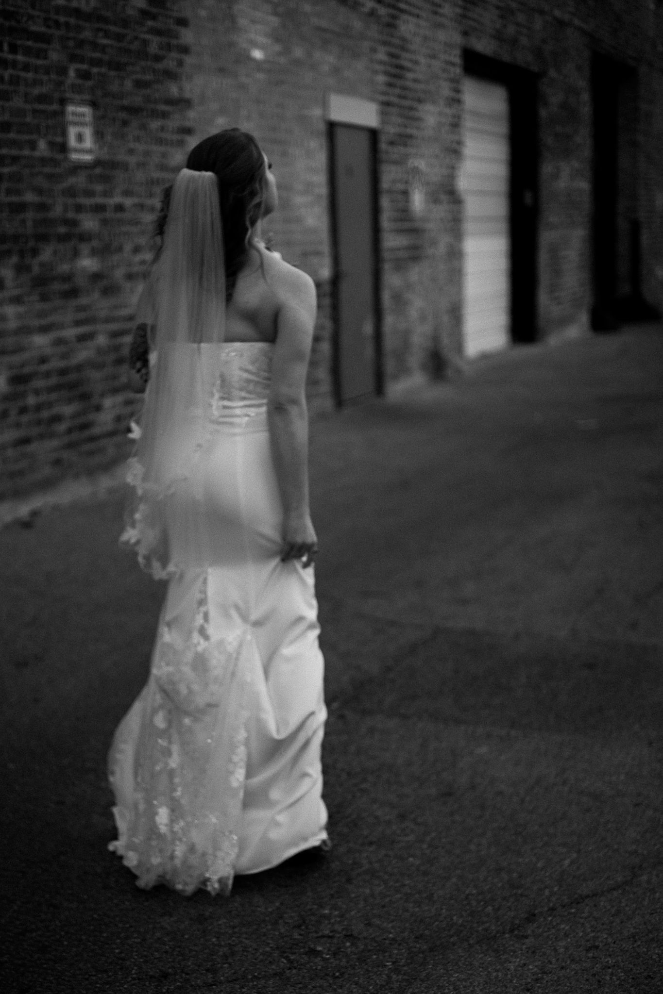 A bride in a flowing white wedding dress walks away down a dark brick alley in black and white photography.