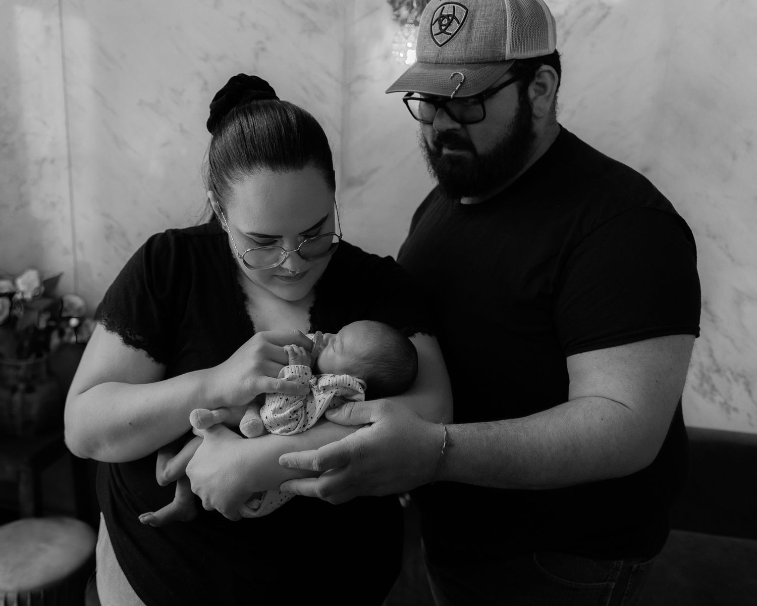 A black and white photograph shows two adults tenderly holding and looking down at a newborn baby.