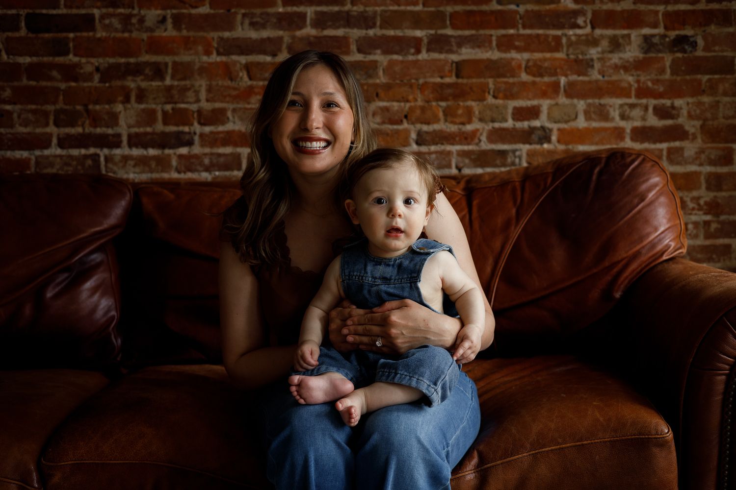 A smiling woman sits on a brown leather couch holding a cute baby in denim overalls against a brick wall.