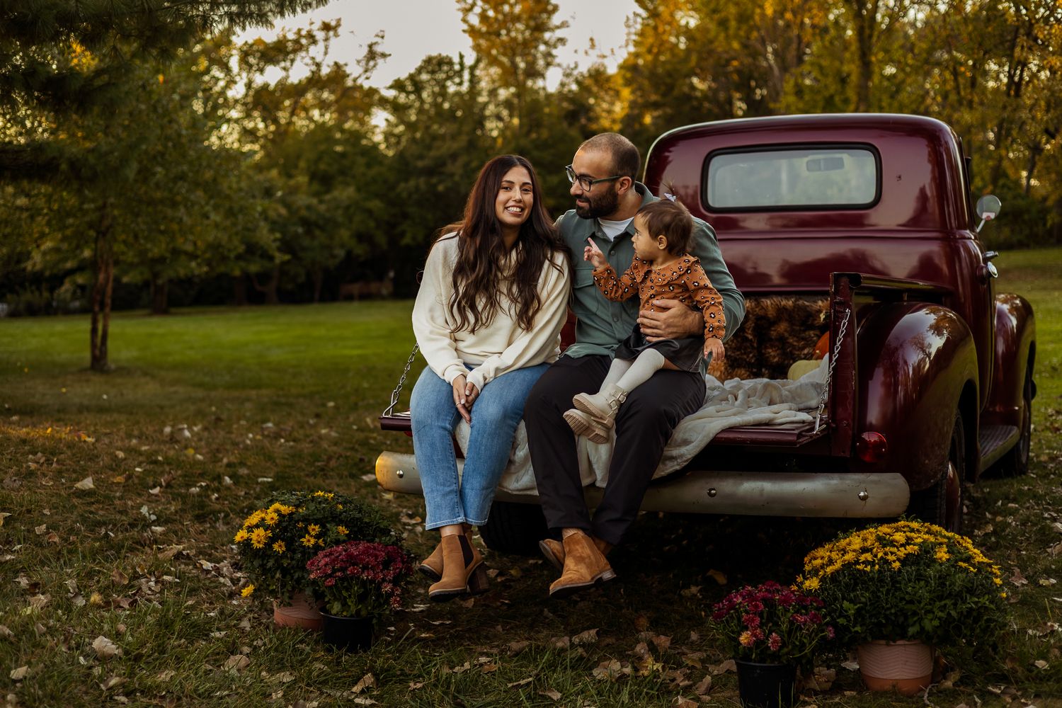 A family enjoys a fall afternoon together near a vintage red pickup truck surrounded by autumn mums and foliage.
