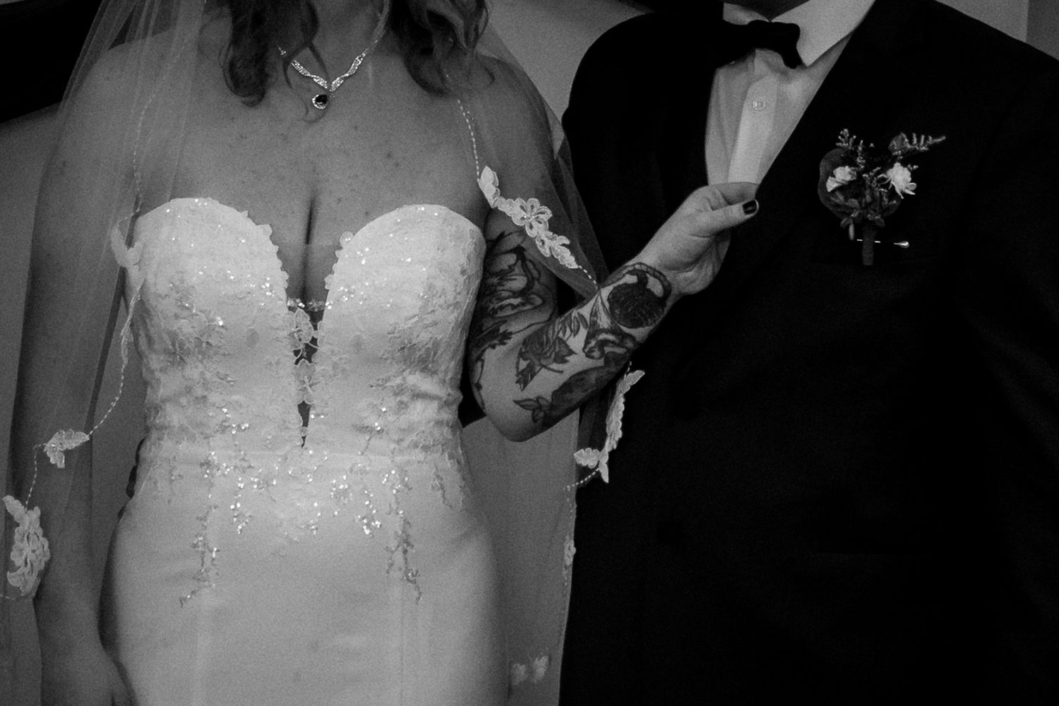 Black and white wedding photo showing close-up detail of bride's lace dress and groom's tuxedo with boutonniere.