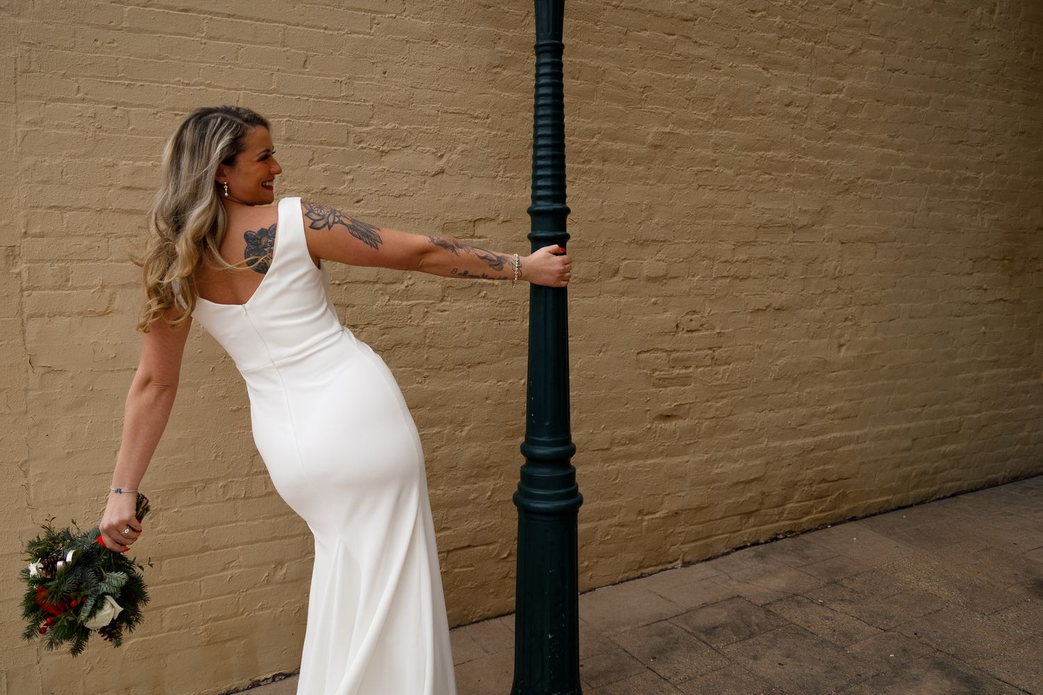 A person in a fitted white wedding dress holds a bouquet while leaning against a green lamppost against a beige wall.