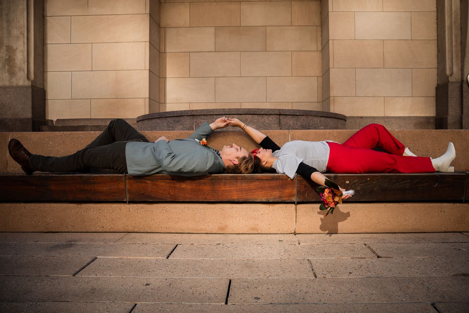 Two people lying close together on a wooden bench in an urban setting with stone walls.