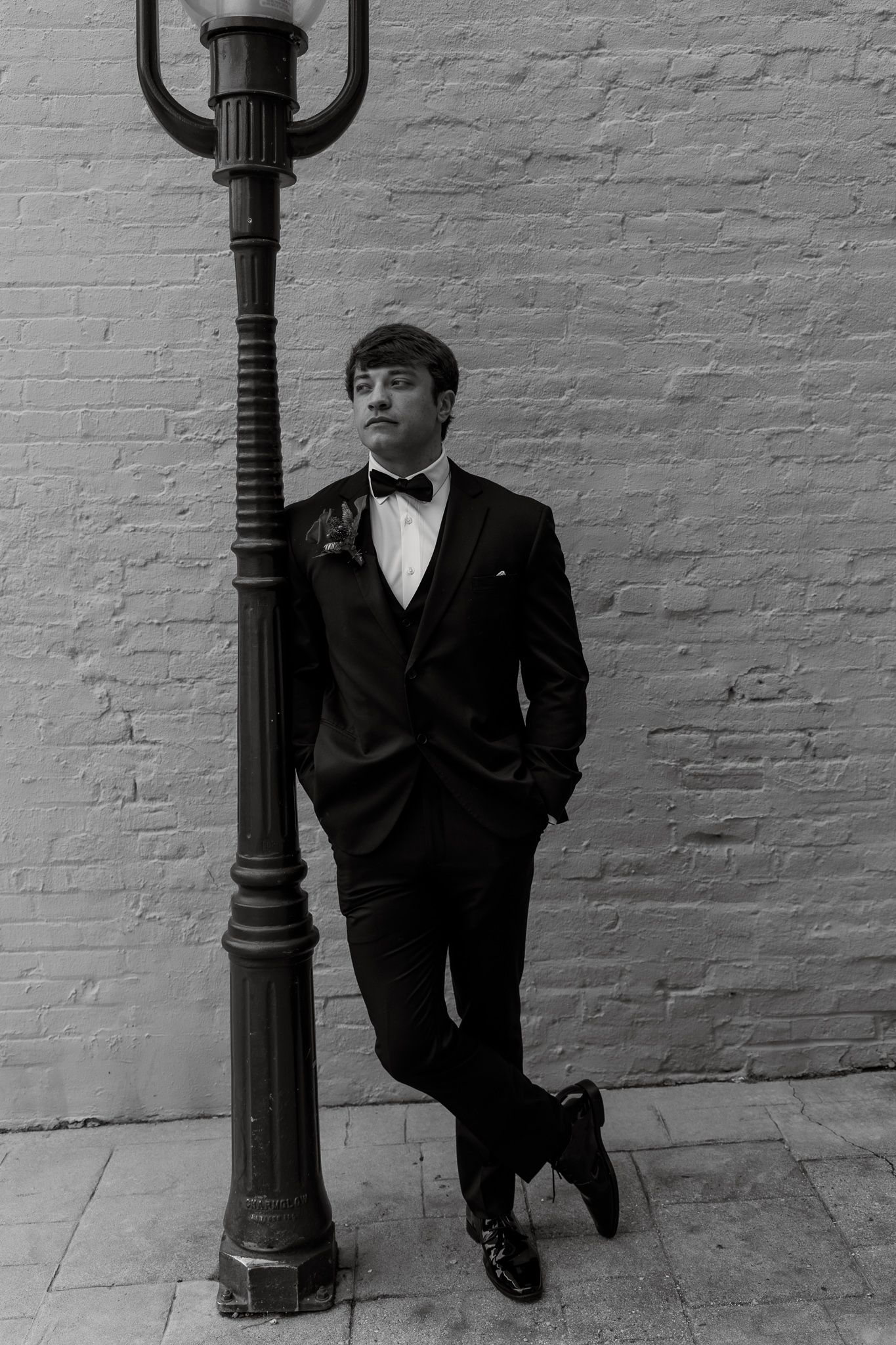 Black and white portrait of a person in a tuxedo and bow tie leaning against a vintage street lamp post.