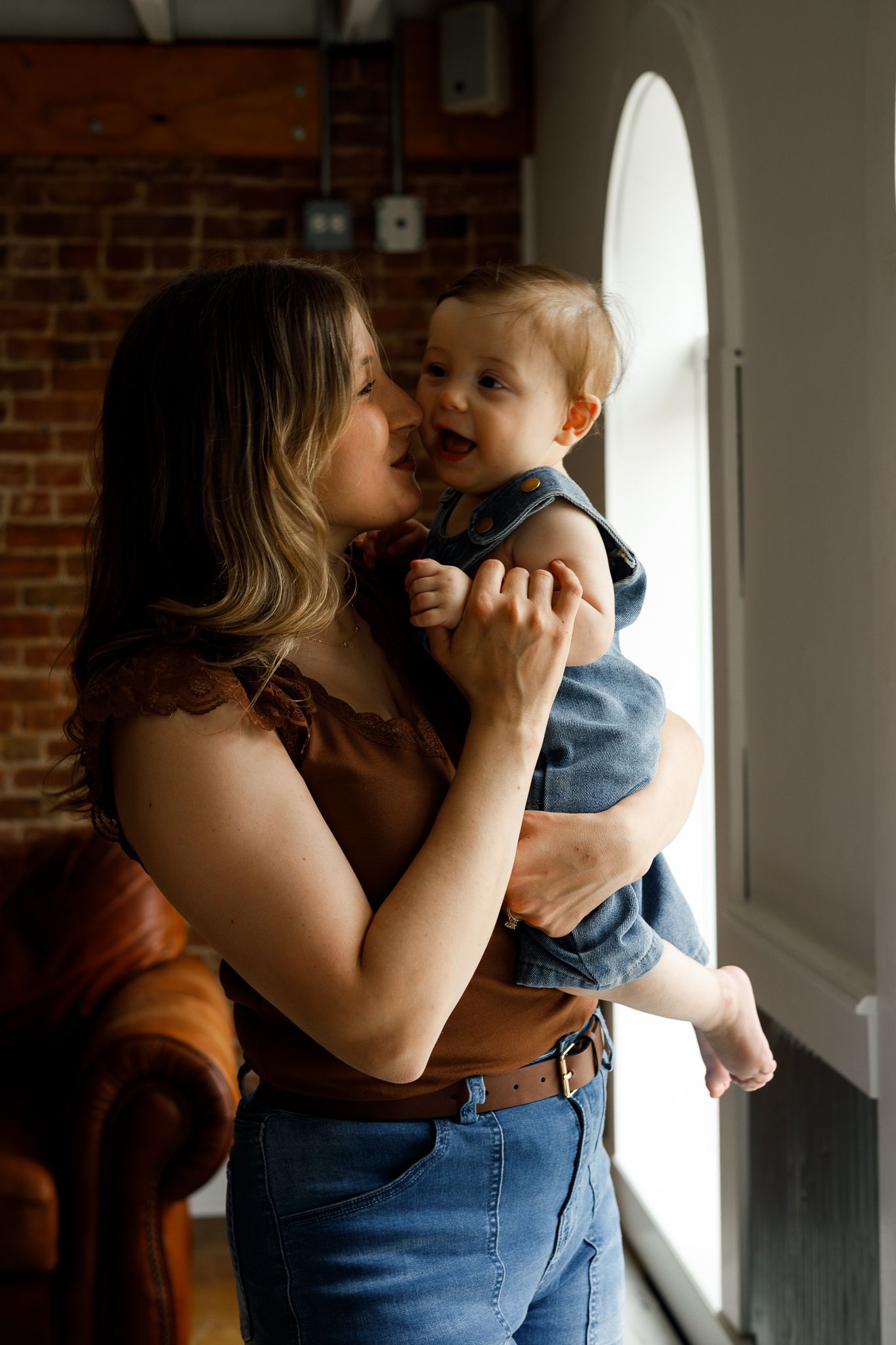 A young mother holds her smiling baby near a window in a brick-walled room, sharing a tender moment.