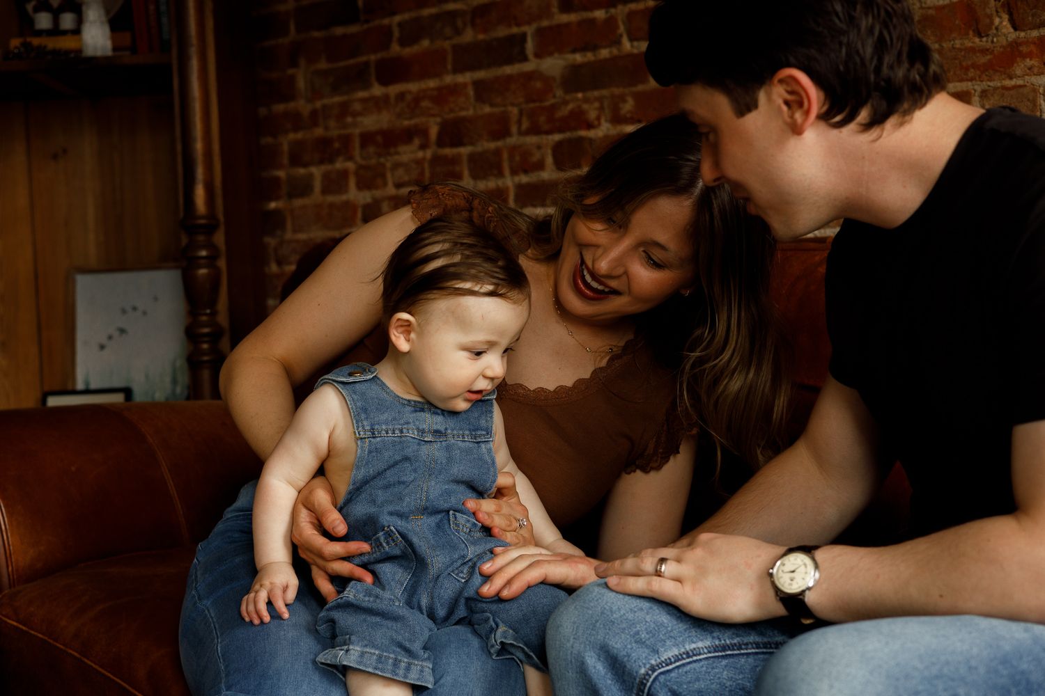 Parents smiling at their baby in denim overalls on a leather couch with brick wall background.