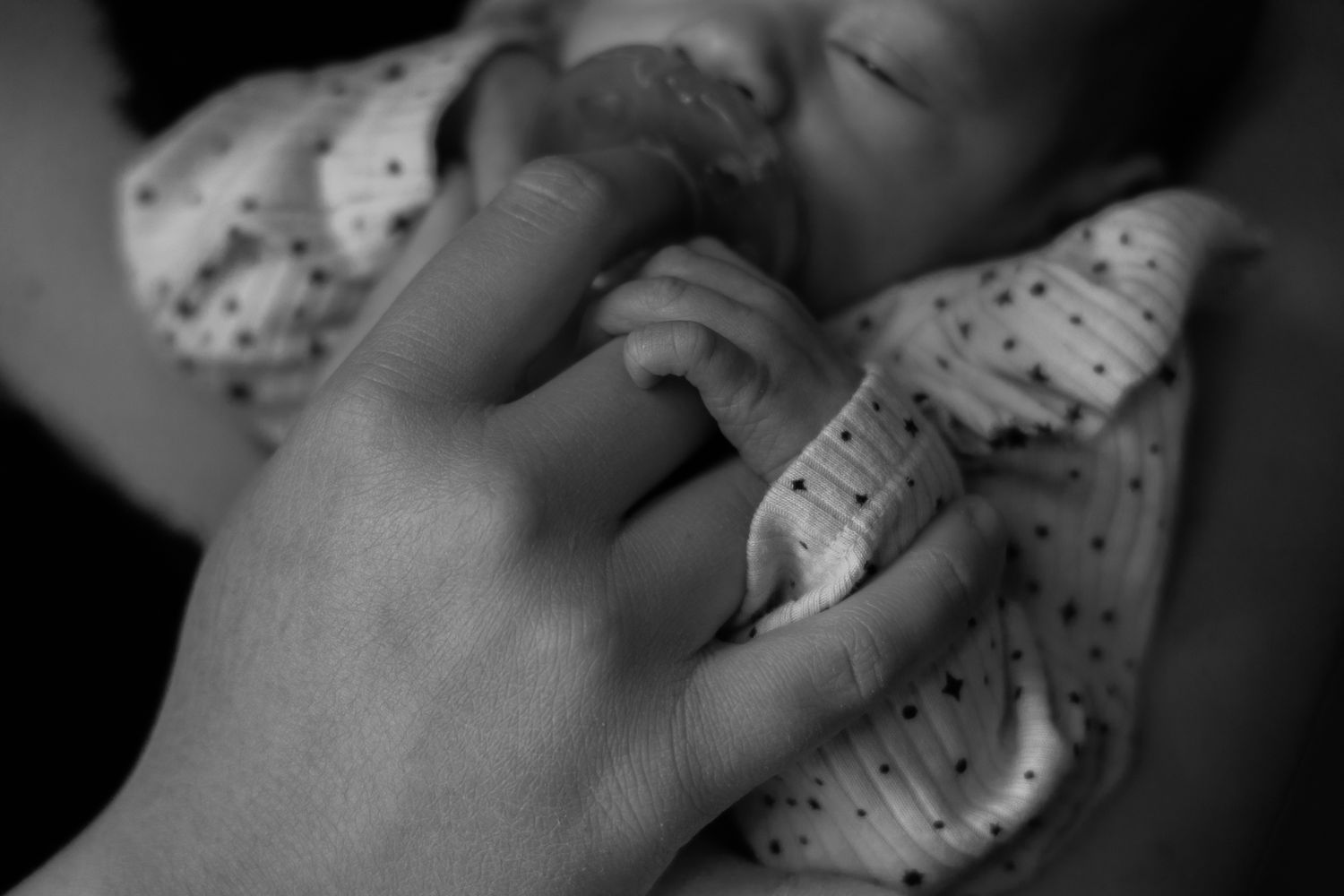 A close-up black and white image of a small hand grasping an adult's finger.
