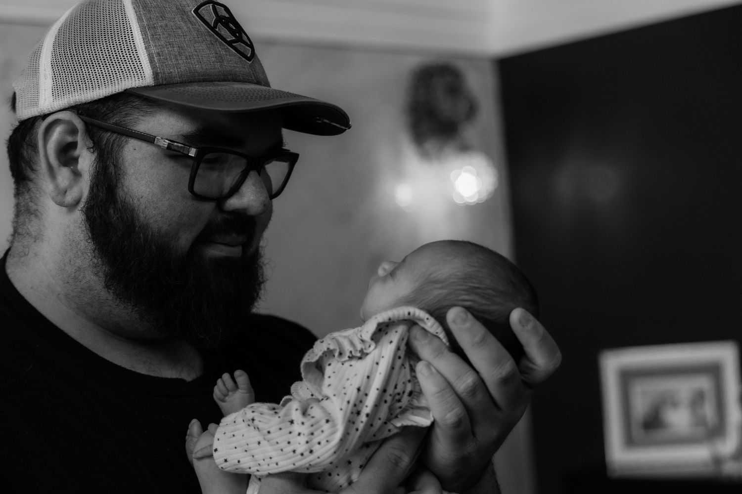 A bearded man wearing glasses and a baseball cap holds a small baby in a tender black and white moment.