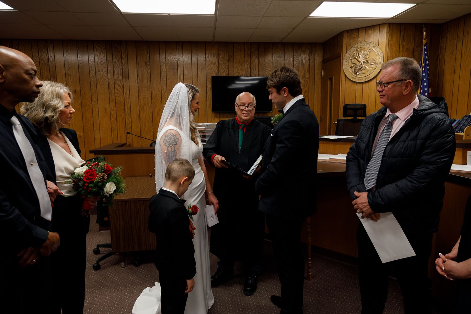 A wedding ceremony takes place in a wood-paneled courtroom with an officiant presiding over the proceedings.