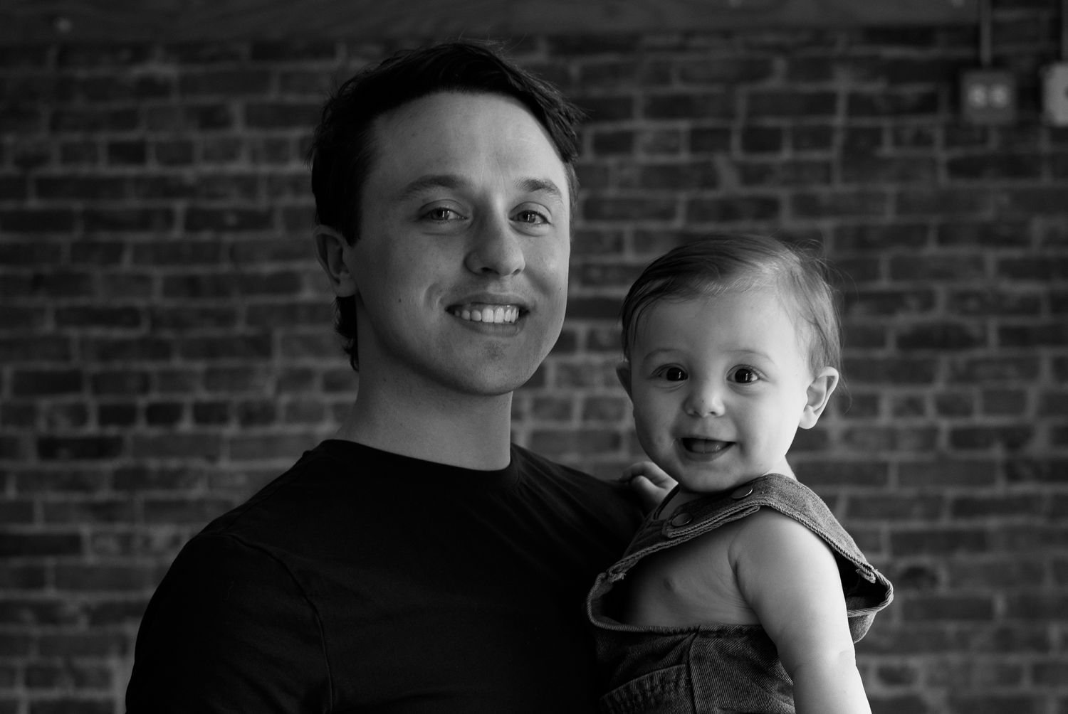Black and white photo of a smiling young man holding a happy toddler in front of a brick wall.