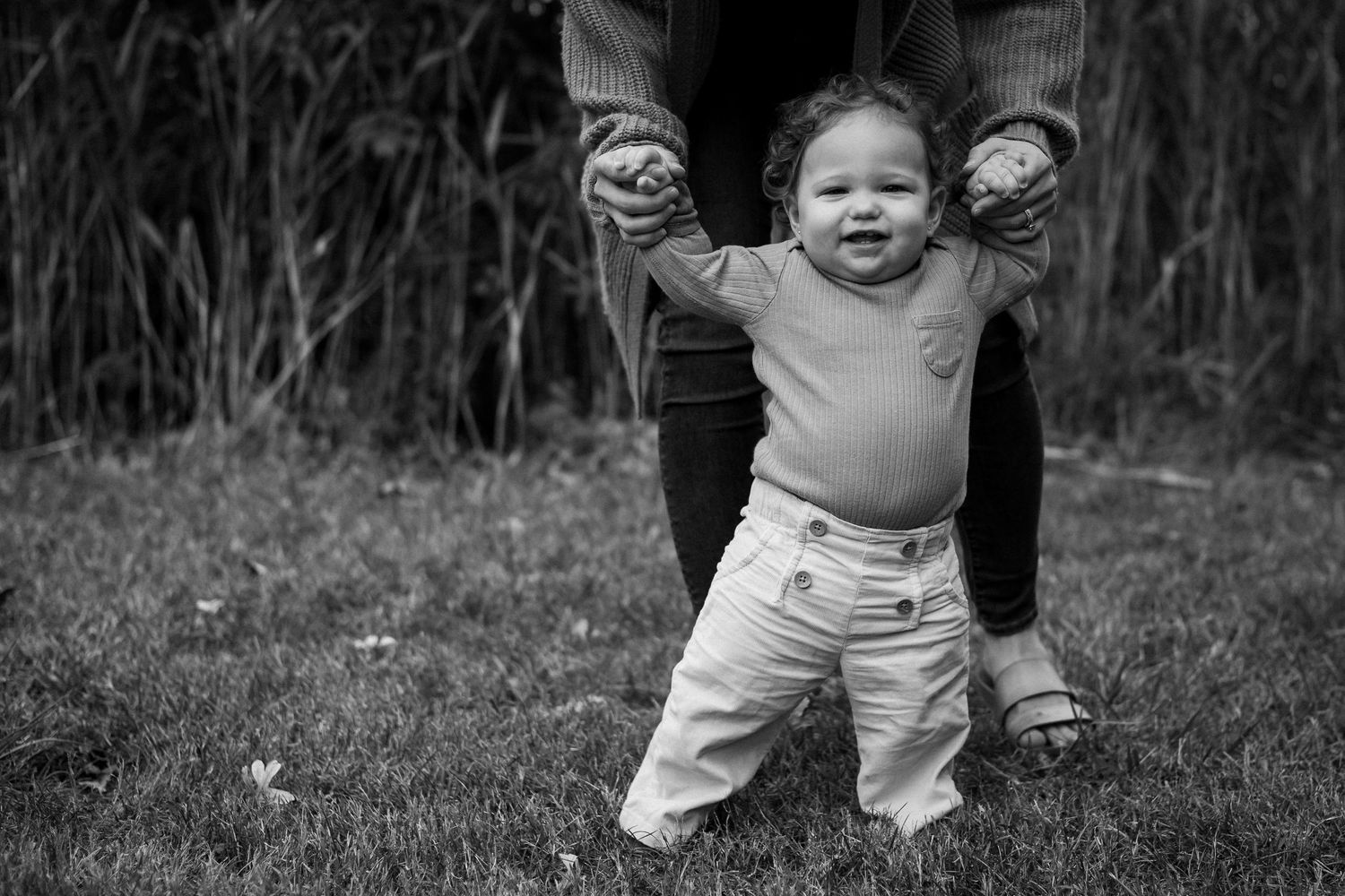 Black and white photo series showing a toddler learning to walk outdoors in grass and natural surroundings.