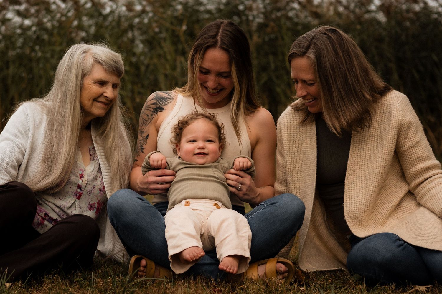 A multi-generational family sitting together outdoors on grass with autumn foliage in the background.