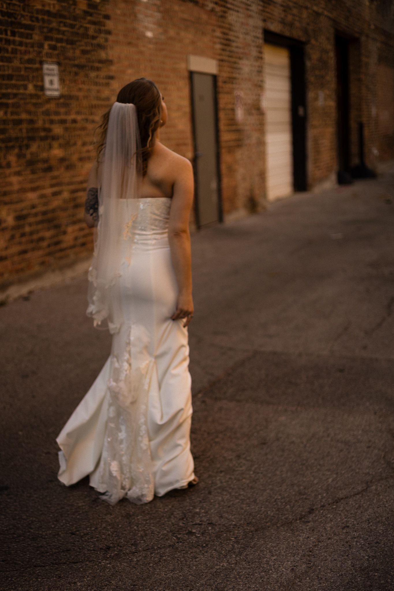A bride in a white strapless wedding gown and veil stands in an alley against rustic brick walls.