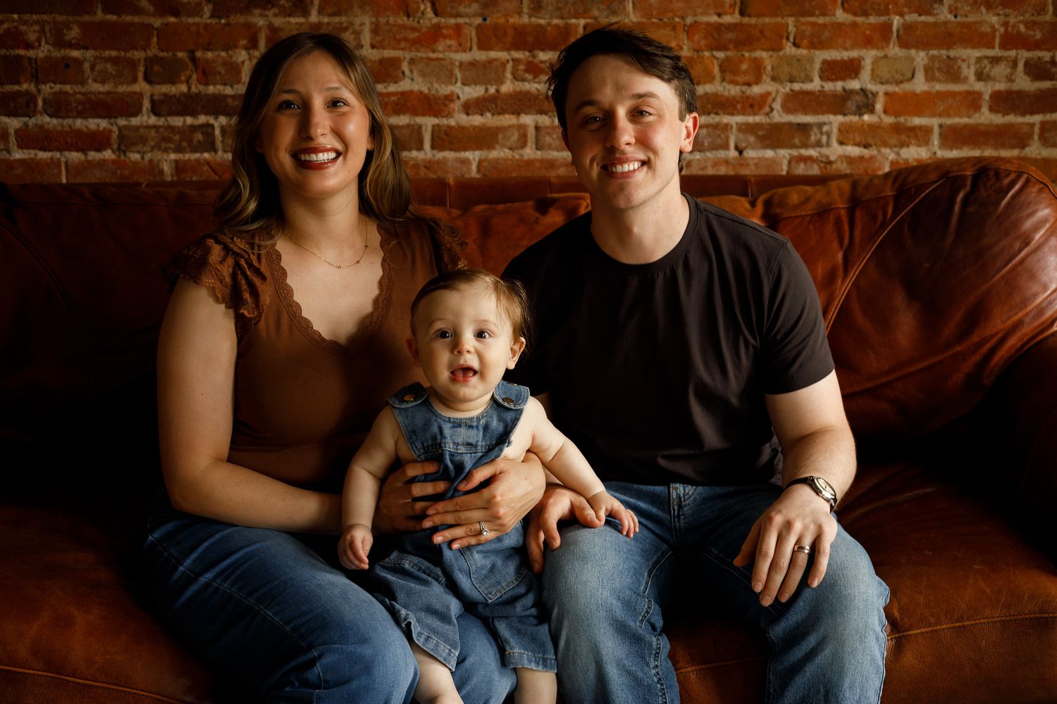 Happy young family with baby in denim overalls posing on leather sofa against brick wall.