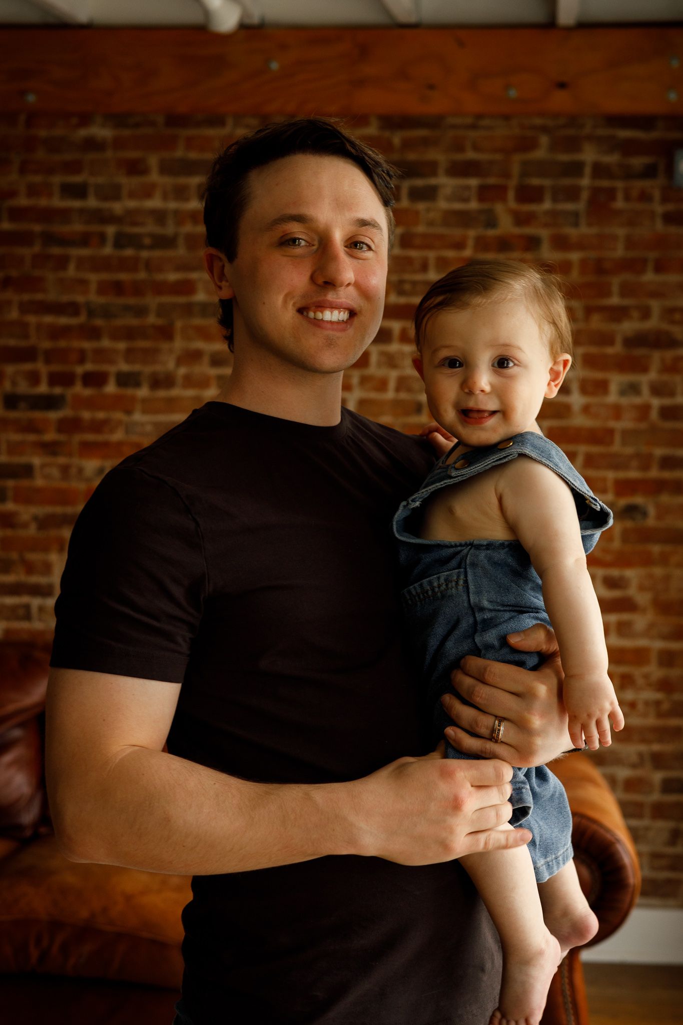 Smiling man in black t-shirt holding a happy baby in denim overalls in front of a brick wall.