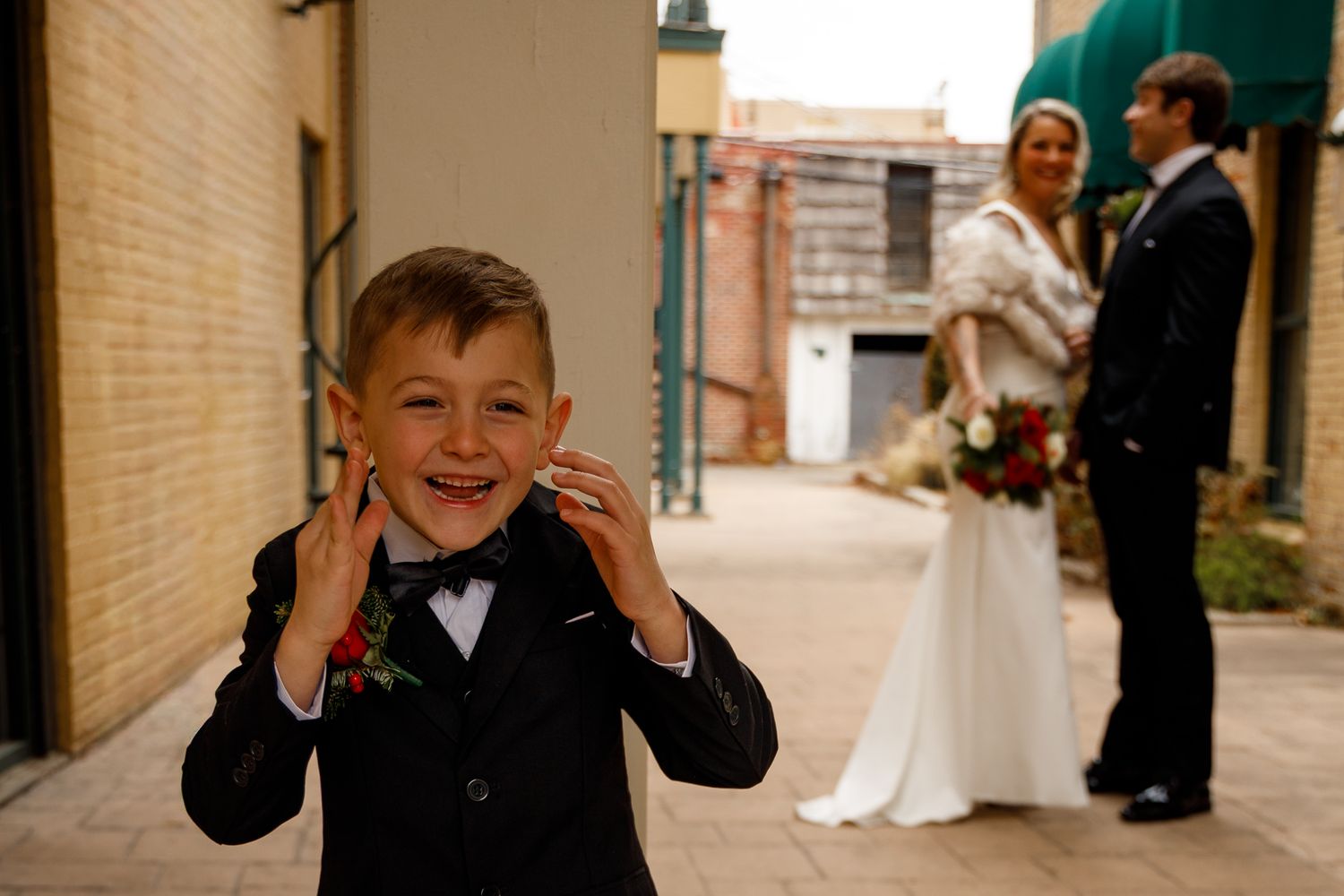 A joyful young ring bearer in a black tuxedo makes a silly face while a wedding couple stands in the background.