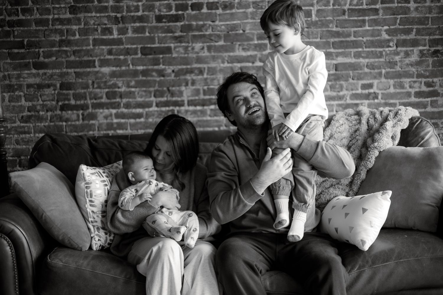 Family sitting together on couch against exposed brick wall in black and white photograph.