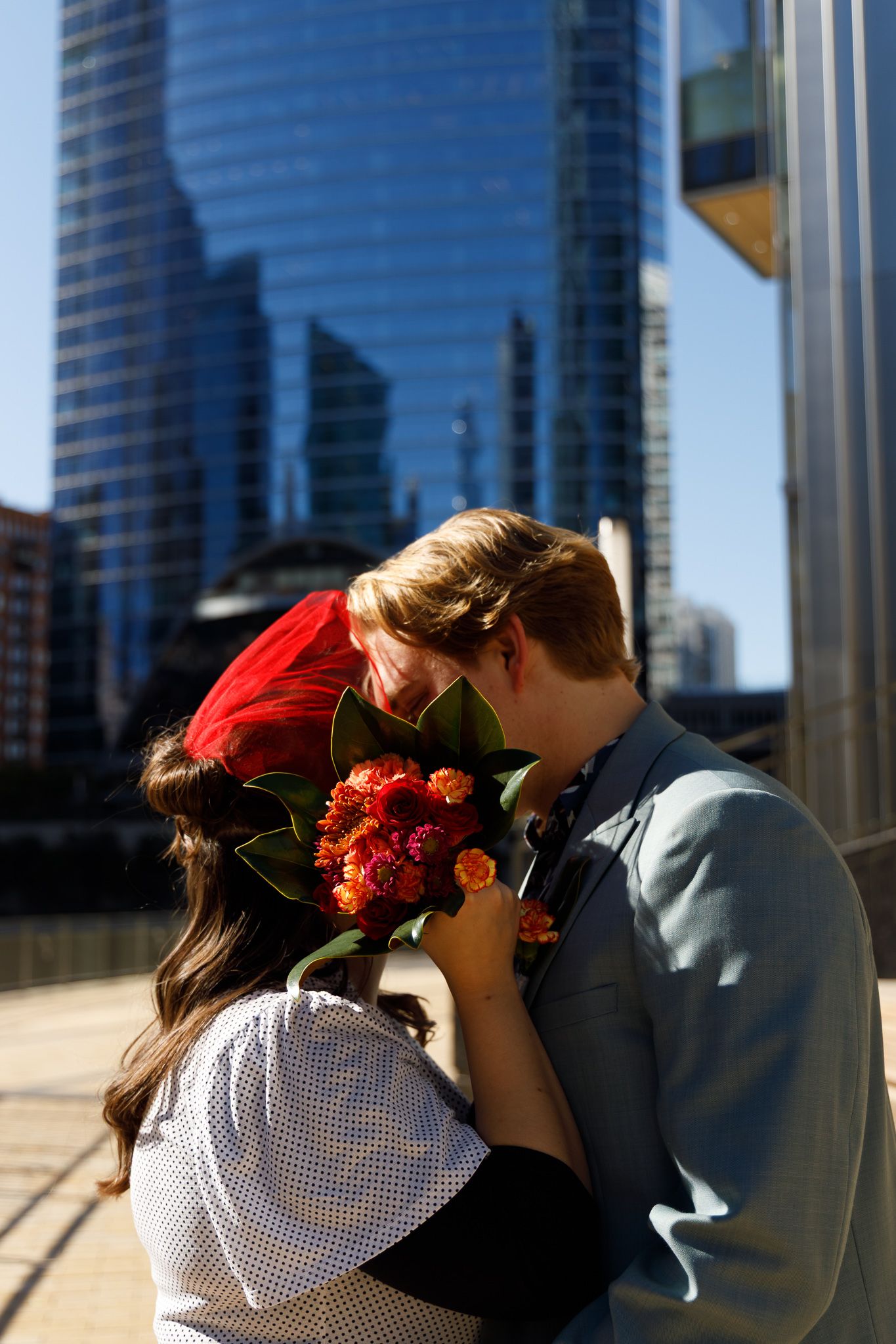 A romantic couple shares a tender moment with orange flowers against a modern city backdrop of glass skyscrapers.