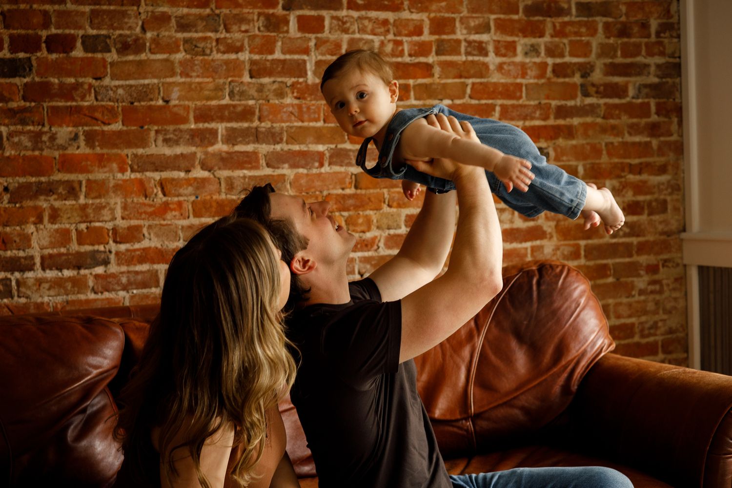 Happy family on brown leather sofa, father lifting smiling toddler in air while mother watches, brick wall background.