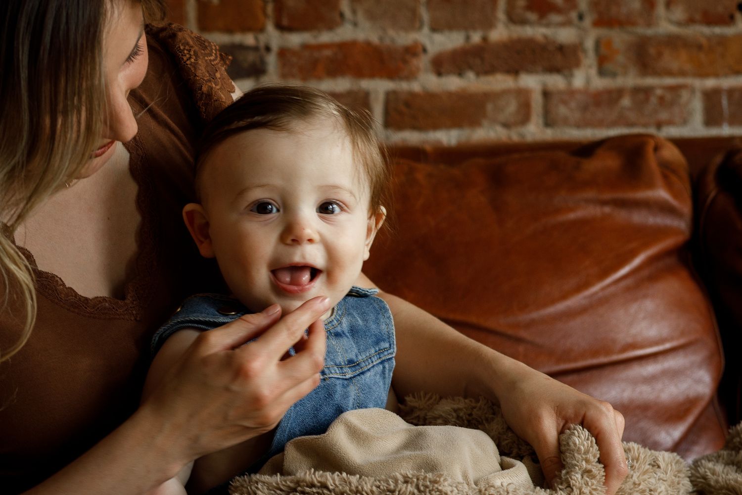 Smiling baby in denim overalls sitting with mother on leather couch near brick wall.