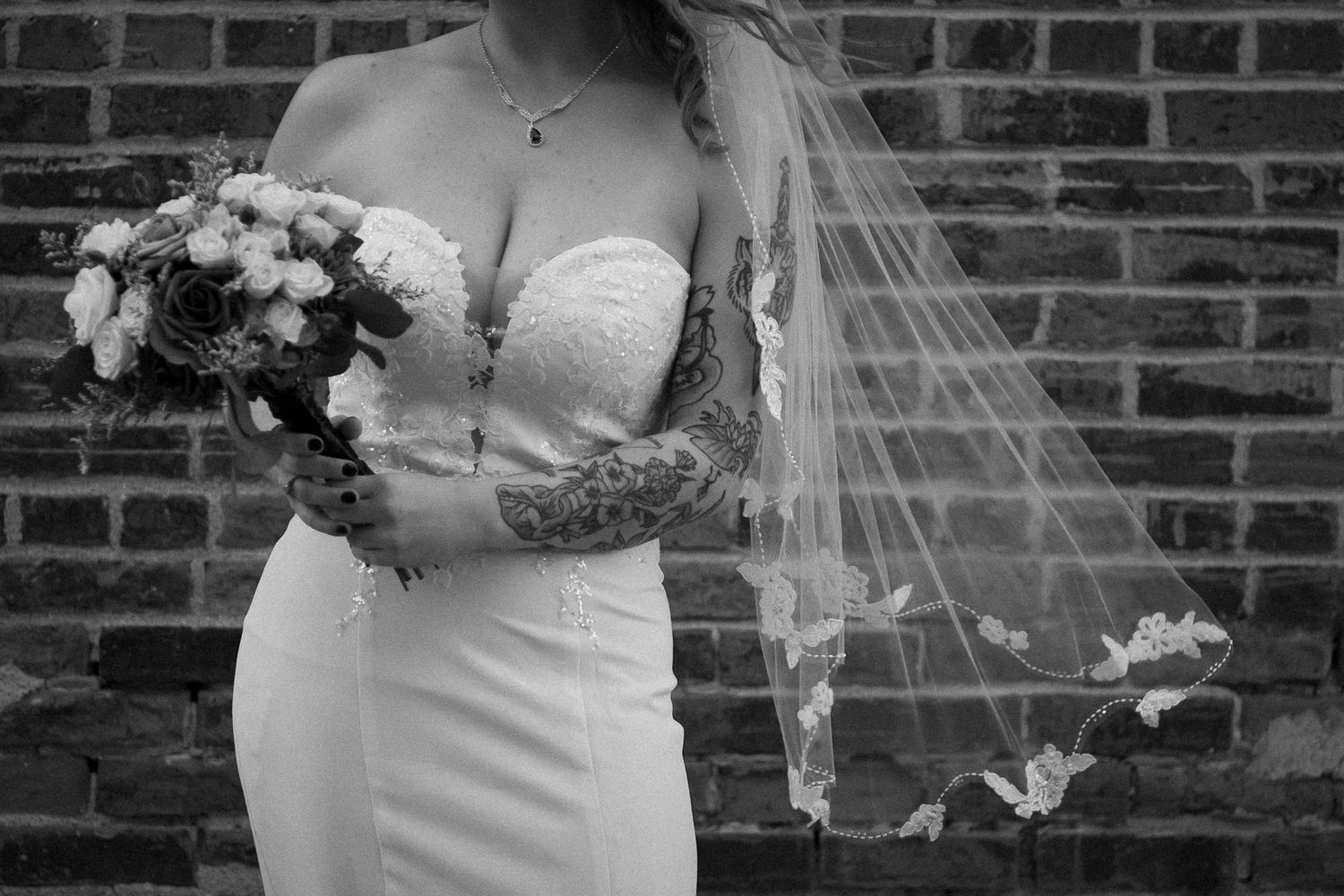A bride in a strapless white wedding gown holds a bouquet while standing against a rustic brick wall.