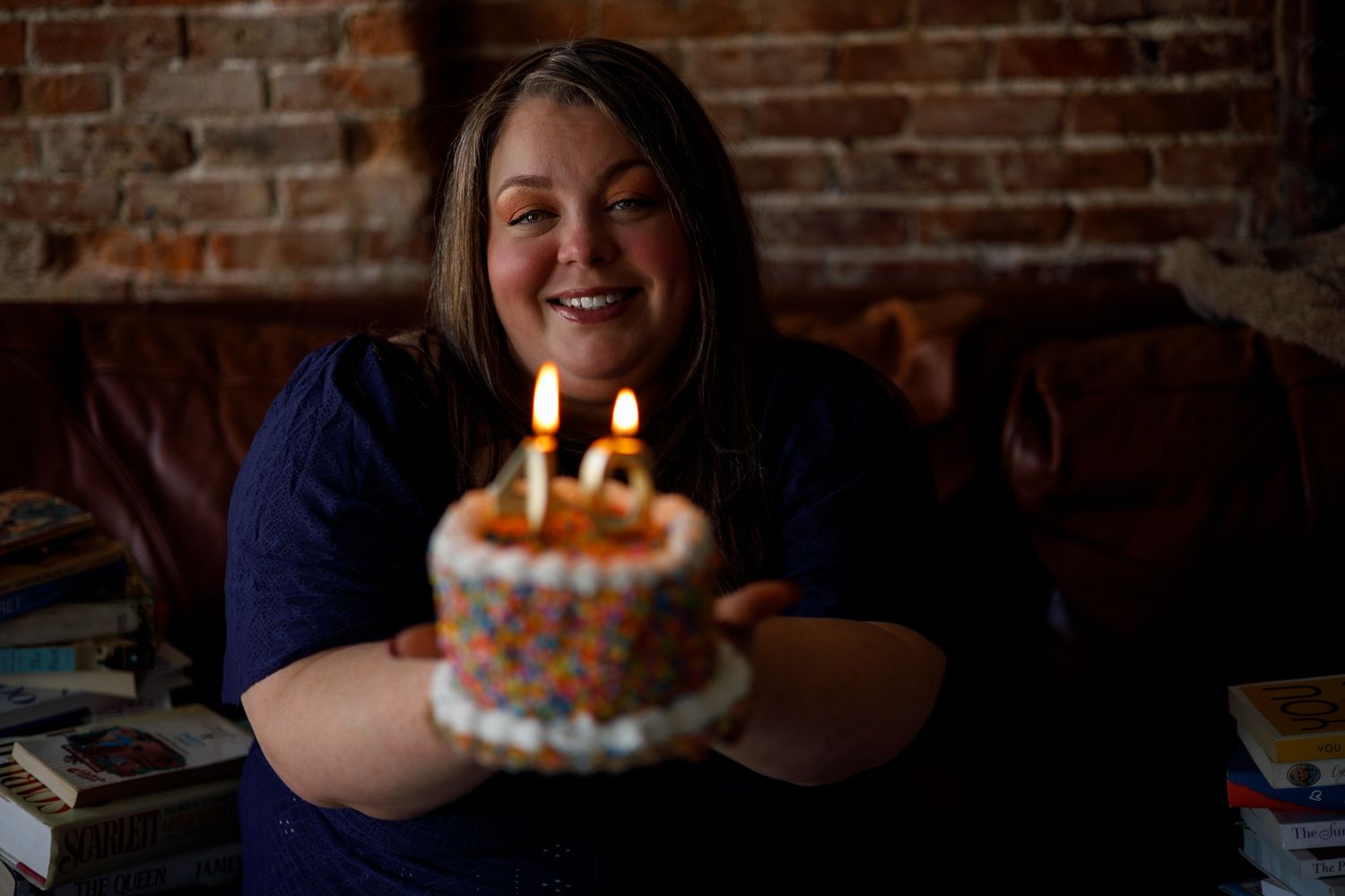 Woman smiling while holding a small decorated birthday cake with lit candles in a cozy brick-walled room.