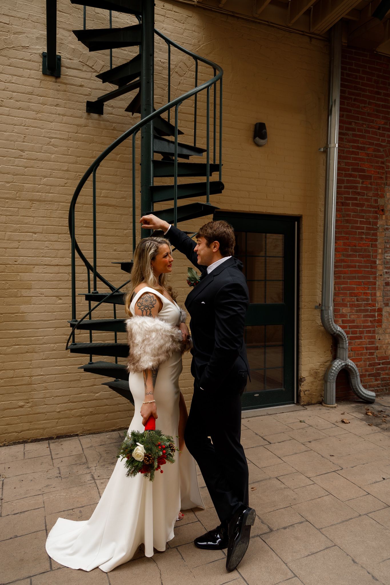 A bride in white dress and groom in black suit share an intimate moment by a spiral staircase.