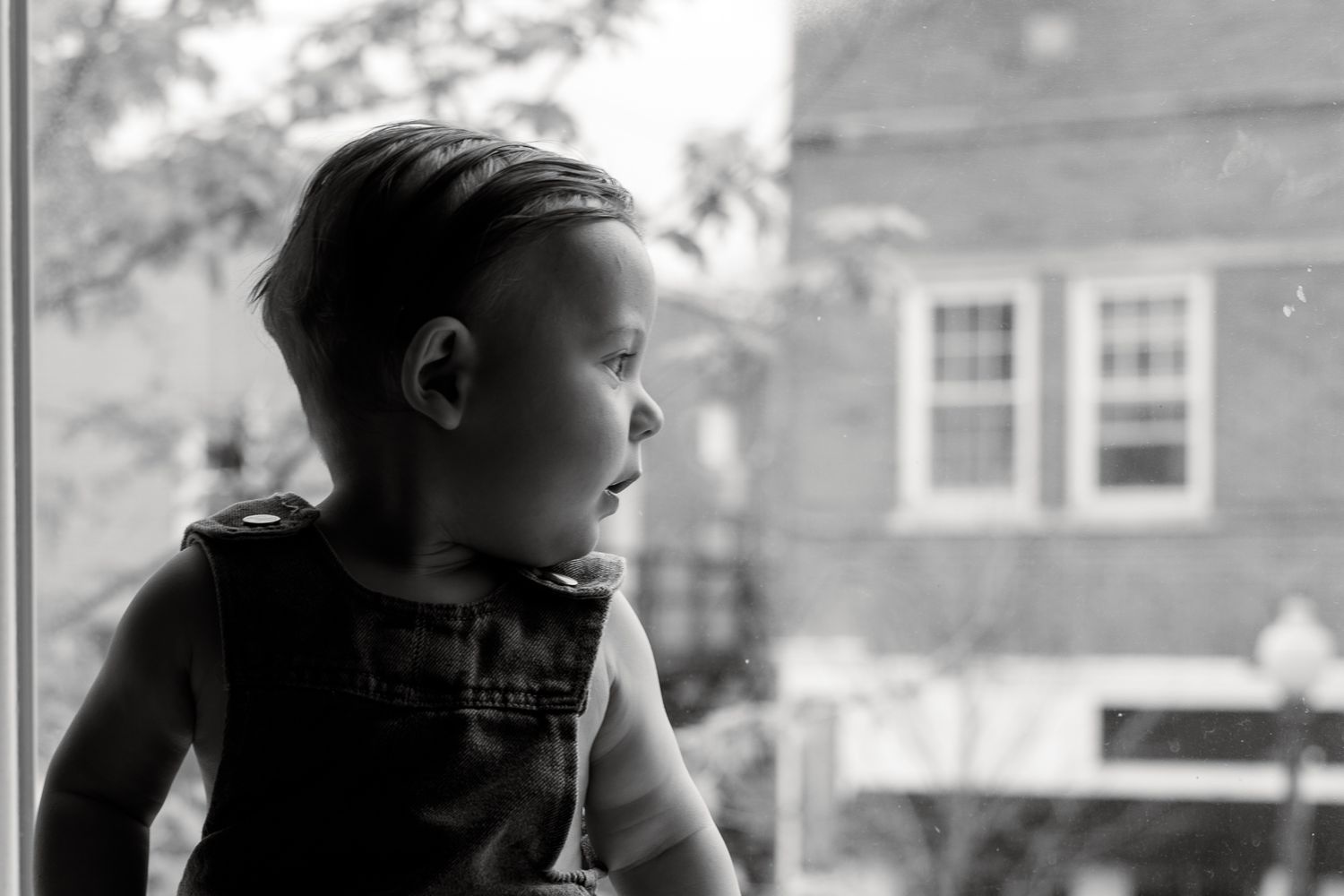 Black and white photo of a young boy in overalls gazing out a window at a brick building outside.