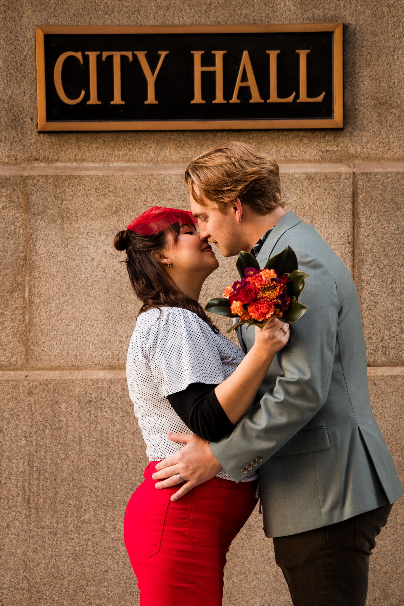 A couple in formal attire share a romantic moment in front of a city hall building.