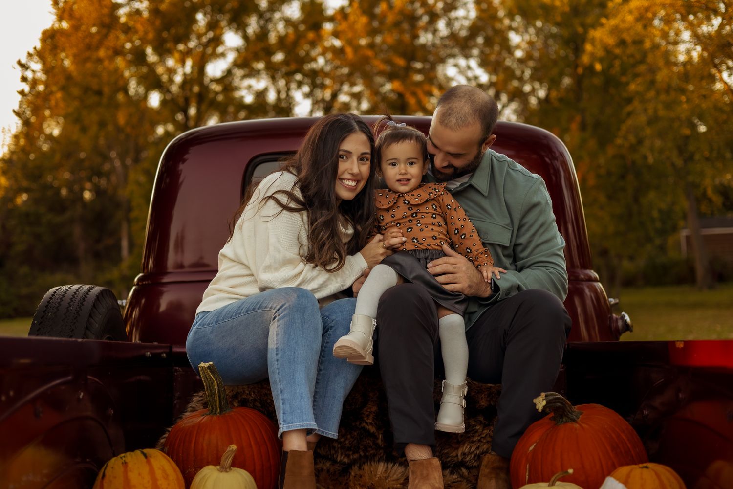 A family sits in the bed of a red pickup truck surrounded by pumpkins during a warm autumn sunset.