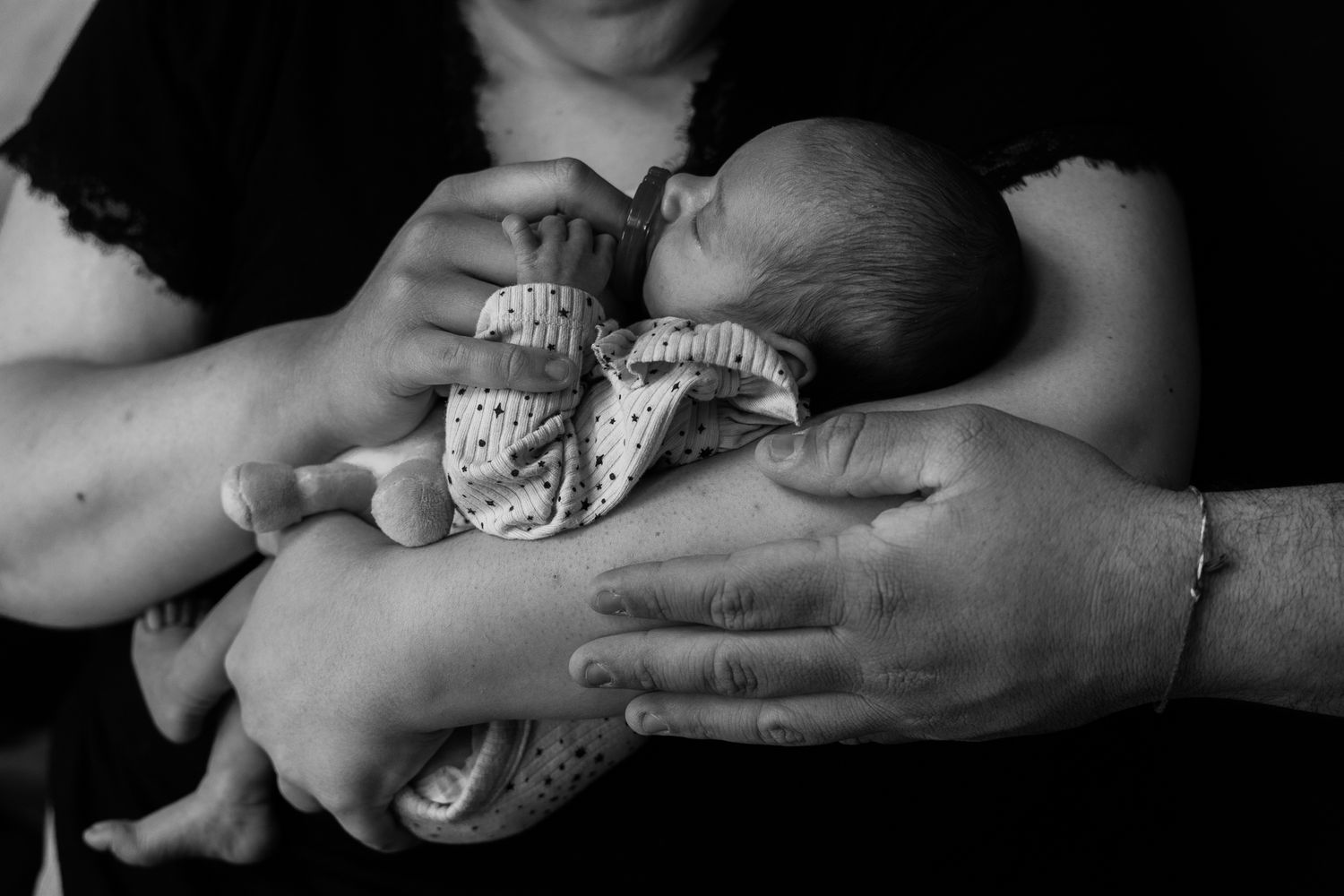 Black and white photo of hands tenderly cradling a newborn baby wrapped in patterned fabric.