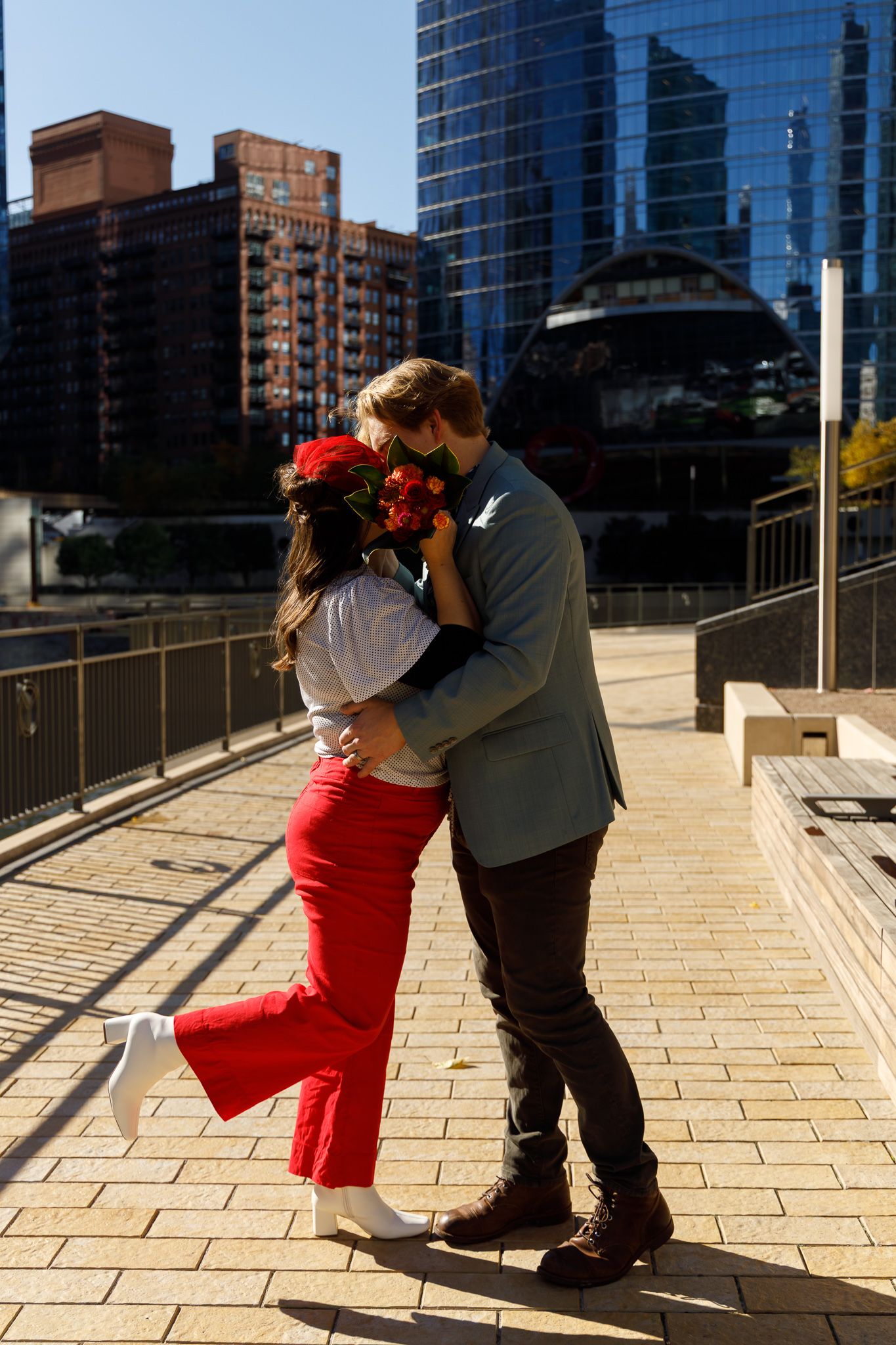 A couple embraces on a sunny urban walkway with red pants and city buildings in the background.