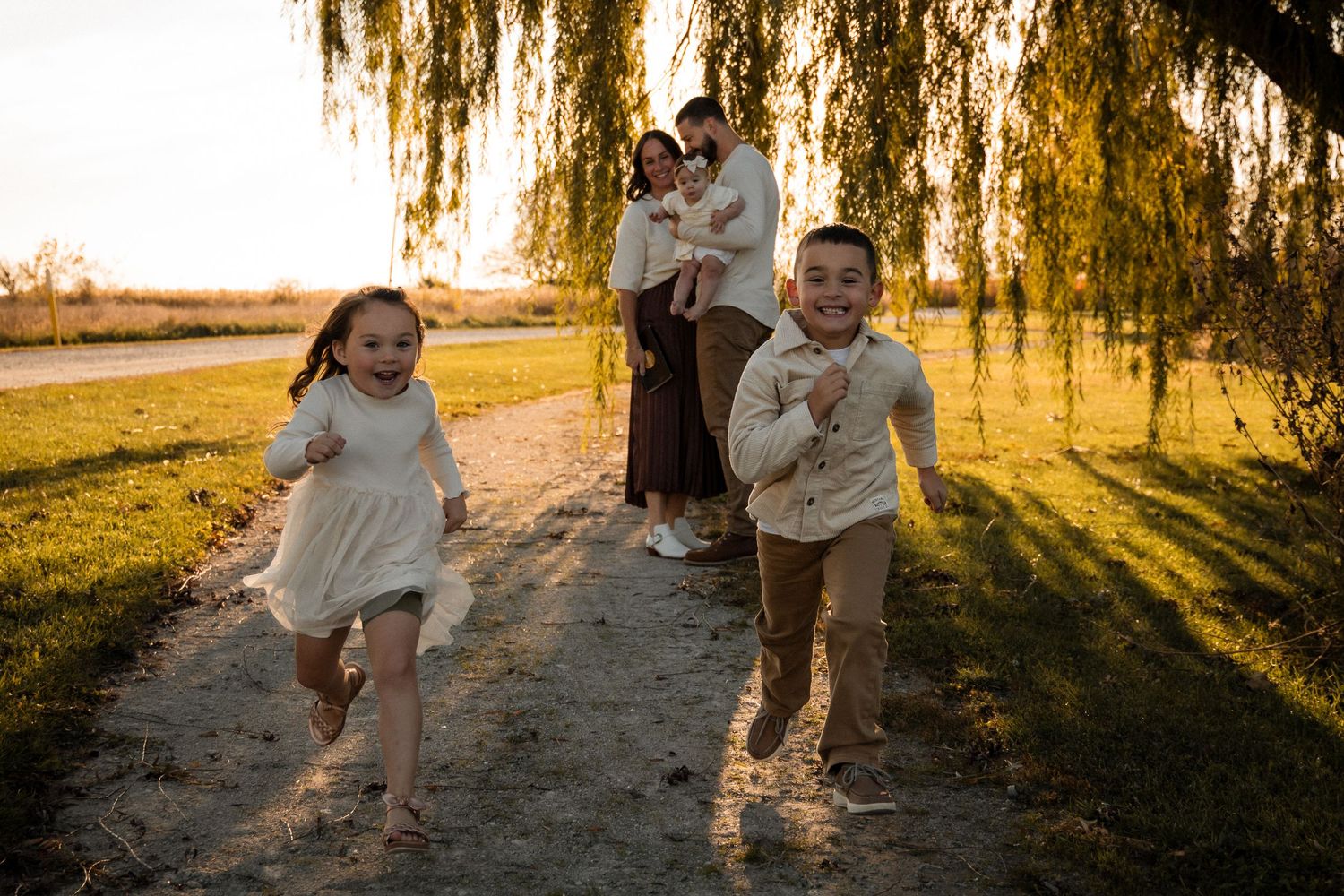 Children running playfully at sunset under a weeping willow tree.