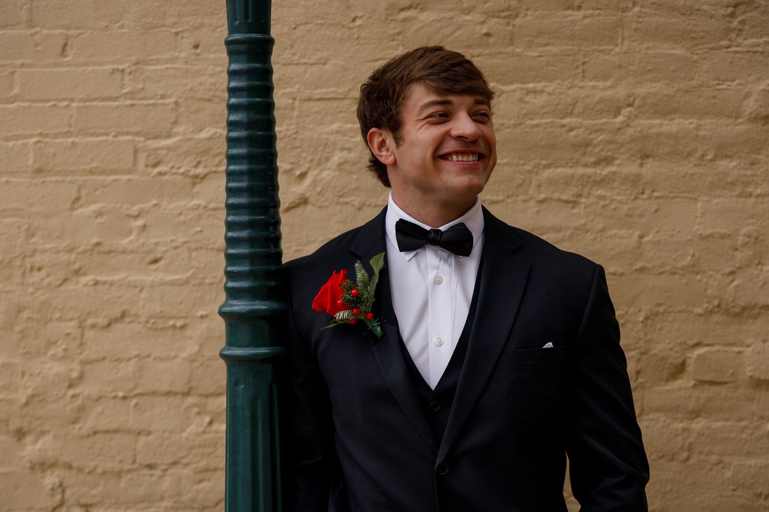 A person in a black tuxedo and bowtie smiles while standing against a beige brick wall with green posts.